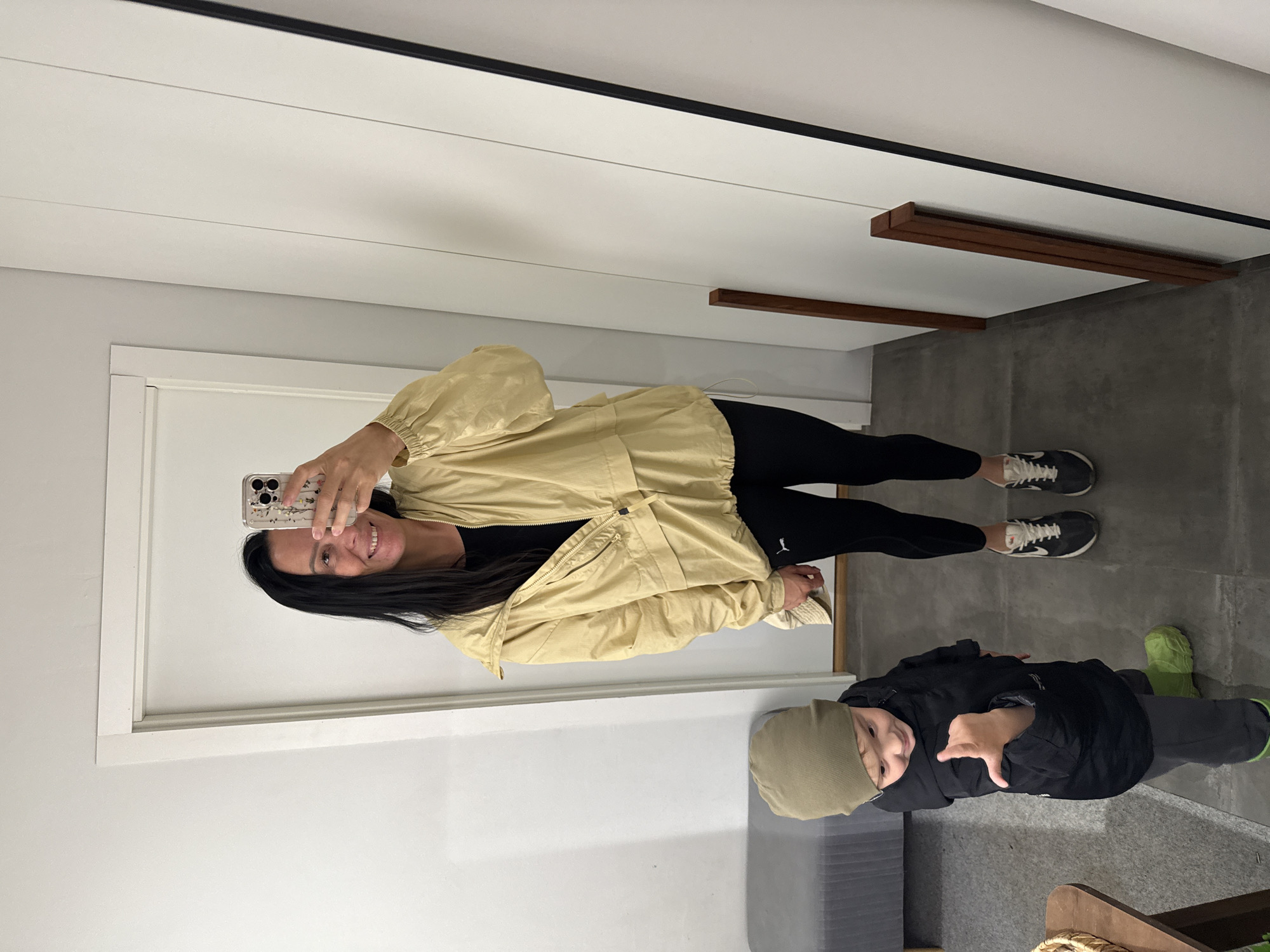 A woman and a small child are standing in a hallway. The woman is wearing a light yellow jacket, black leggings, and sneakers. She is holding a phone in her hand and taking a mirror selfie. The child is wearing a black jacket, gray pants, a green beanie, and green boots. The child is looking down and appears to be talking or gesturing. The hallway has a gray carpeted bench on the left, and white walls. A tall mirror with a black frame is on the right side of the image. The floor is a dark gray tile. The overall scene suggests a domestic or retail setting. There is no indication of the city of Chaikovsky, Russia.