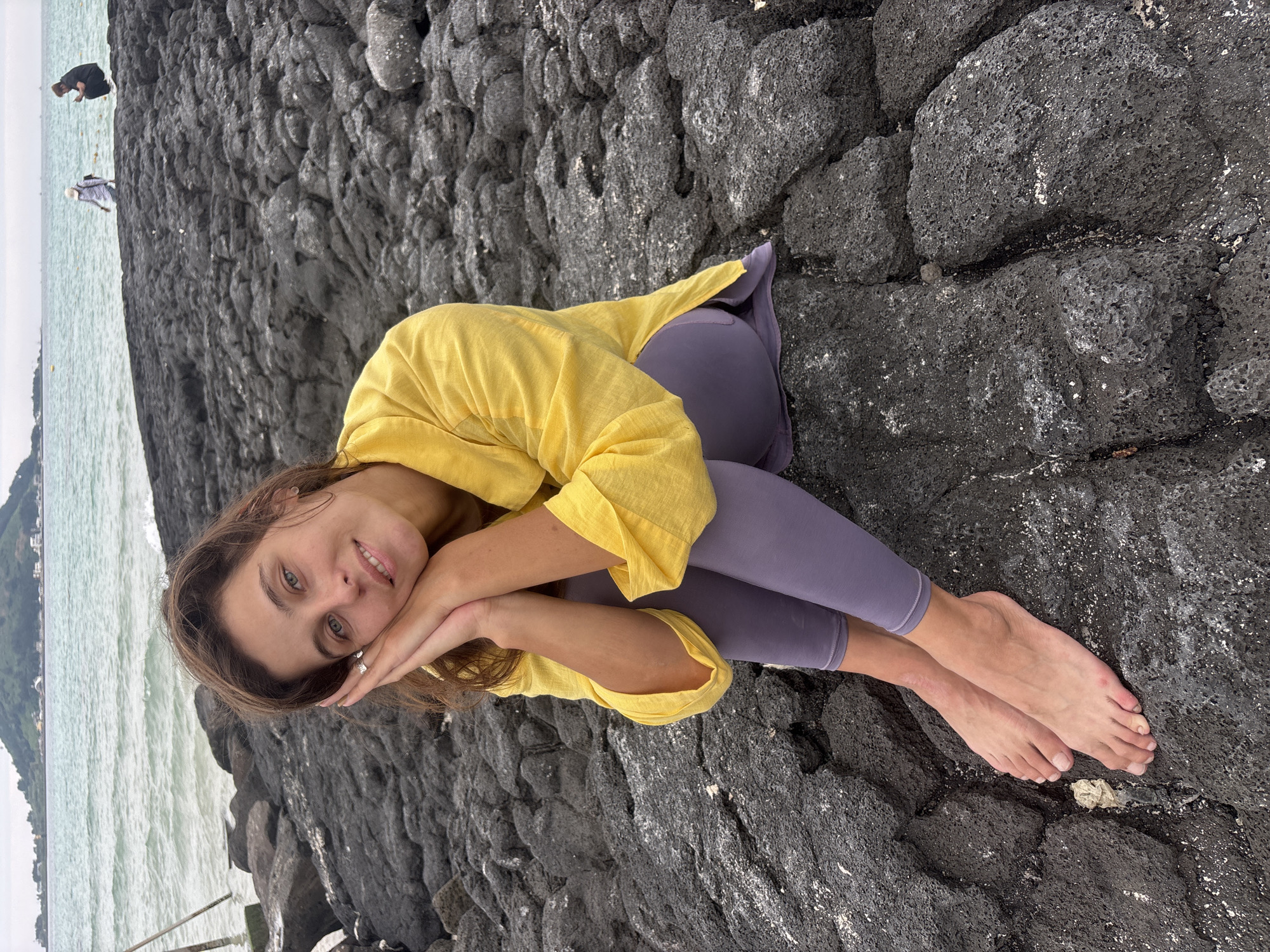 A young woman with long brown hair and blue eyes sits on dark volcanic rocks by the sea. She is wearing a yellow, long-sleeved shirt and purple leggings. Her head is resting on her hands, and she is smiling at the camera. In the background, the ocean waves are visible, and a green, hilly landscape can be seen on the horizon. There are two other people in the distance, one in a black top and another in a white and blue patterned top, walking on the rocks. The sky is overcast, suggesting a cloudy or potentially rainy day. The setting appears to be a coastal area with distinctive dark, porous rock formations, characteristic of volcanic landscapes found on Jeju Island, South Korea. The woman's pose suggests a relaxed and contemplative mood. No visible text is present in the image.