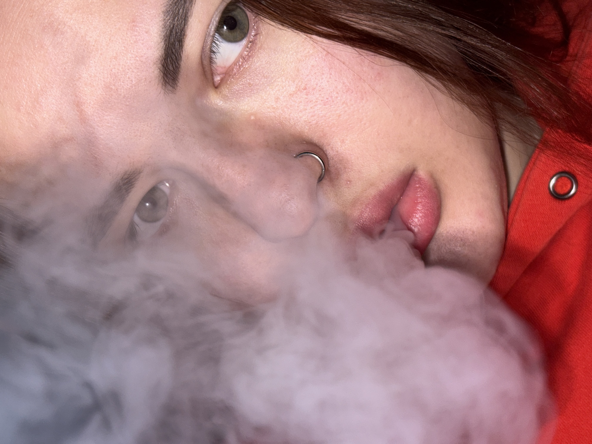 A close-up, angled portrait shows a person with fair skin, brown hair, and light-colored eyes. A silver nose ring is visible on the left nostril. The person's lips are slightly parted. Smoke, or vapor, obscures the lower portion of the face. The person is wearing a red garment, possibly a hooded sweatshirt, as indicated by a visible grommet. The location, Saratovskaya, Russia, is not discernible from the image.