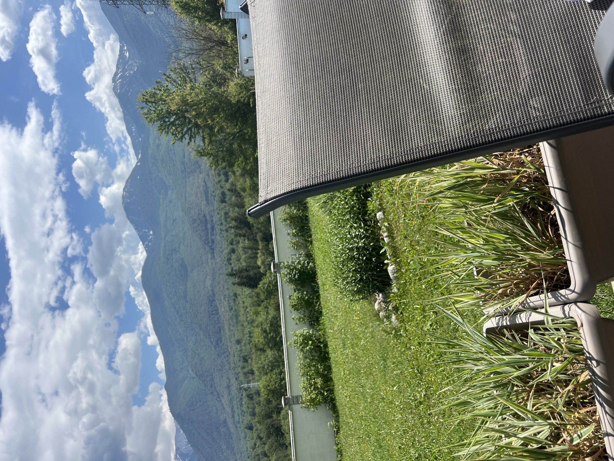 This is a landscape photograph taken in Razlog, Bulgaria, likely on a bright, sunny day with scattered clouds. The image is oriented vertically. In the foreground, a portion of an outdoor furniture item, possibly a patio chair or sun lounger, is visible, its textured fabric taking up the top right section of the frame. Below the furniture, planters filled with lush green and variegated ornamental grass are prominent.
The middle ground features a well-maintained lawn and a white fence that runs horizontally across the frame, separating the manicured area from the natural landscape beyond. Beyond the fence, a dense, green forest covers the lower slopes of a majestic mountain range. The mountains themselves are a prominent subject, their peaks partially obscured by haze and distance, with some snow visible on higher elevations. The sky is a vibrant blue, punctuated by fluffy white clouds, suggesting fair weather.
The scene evokes a sense of peacefulness and natural beauty, with a clear contrast between the man-made elements of the patio and the wild grandeur of the mountains. The lighting suggests it's daytime, likely late morning or early afternoon, given the bright sunlight and clear skies. There is no visible text in the image.
