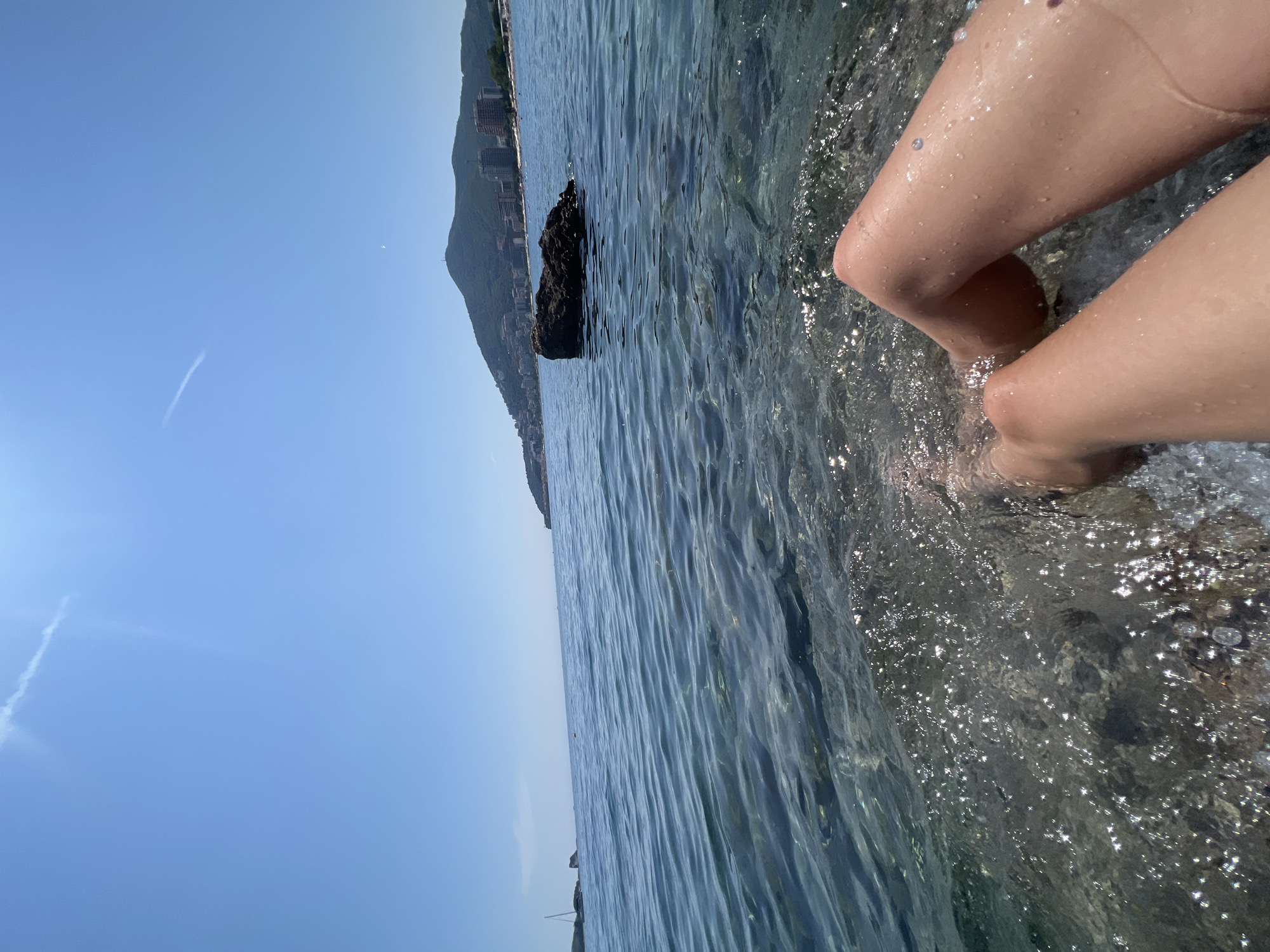 A low-angle, wide shot captures a section of the sea in front of Budva, Montenegro, on a sunny day. The foreground features a person's bare legs submerged in clear, shallow water. The water laps against rocks visible beneath the surface, with sunlight glinting off the waves. In the mid-ground, a dark, textured rock juts out of the water. Beyond this, the expansive blue sea stretches to the horizon, meeting a clear, pale blue sky. In the distance, a hilly landscape rises, dotted with buildings and structures that indicate the presence of the city of Budva. The coastline is visible, with a beach and more buildings lining the shore. The overall scene conveys a sense of relaxation and a summer day by the sea.
