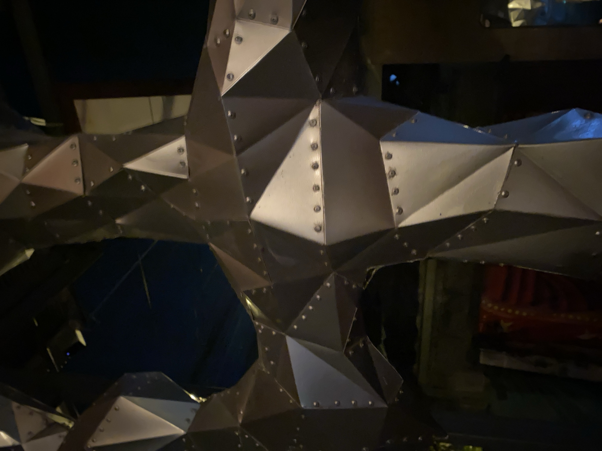 The image shows a close-up of a metallic, geometric structure composed of triangular panels connected with rivets. The panels are arranged to create a multifaceted, three-dimensional form. The setting is dimly lit, with dark areas behind the structure. To the right, a glimpse of a red and patterned object can be seen. The location is ตำบลเกาะพะงัน, Thailand. There are no discernible people or actions within the frame.