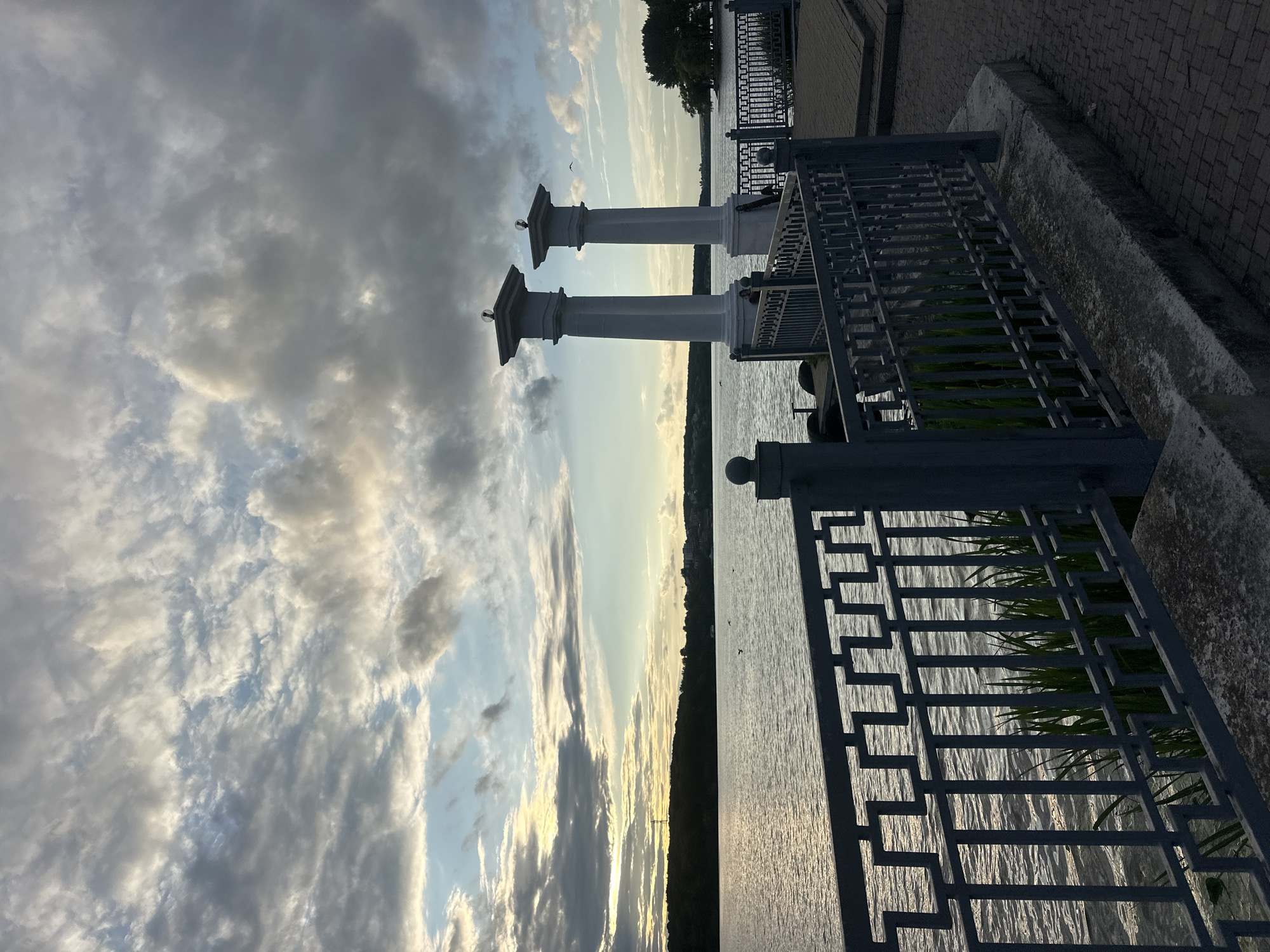 The image depicts a waterfront scene at Ternopil Lake in Ternopil, Ukraine, likely during sunset. Two tall, white classical columns with decorative tops stand prominently on a paved walkway. A grey metal railing with an intricate geometric design separates the walkway from the water. Beyond the railing, the calm lake reflects the soft light of the setting sun, with a treelined shore visible in the distance. The sky is filled with dramatic clouds, showcasing a mix of grey and golden hues, indicating the time of day. There are no people visible in the image, but a few birds can be seen in the sky and on the water, adding a touch of life to the serene landscape. The overall atmosphere is peaceful and scenic, highlighting the natural beauty and architectural elements of the location.
