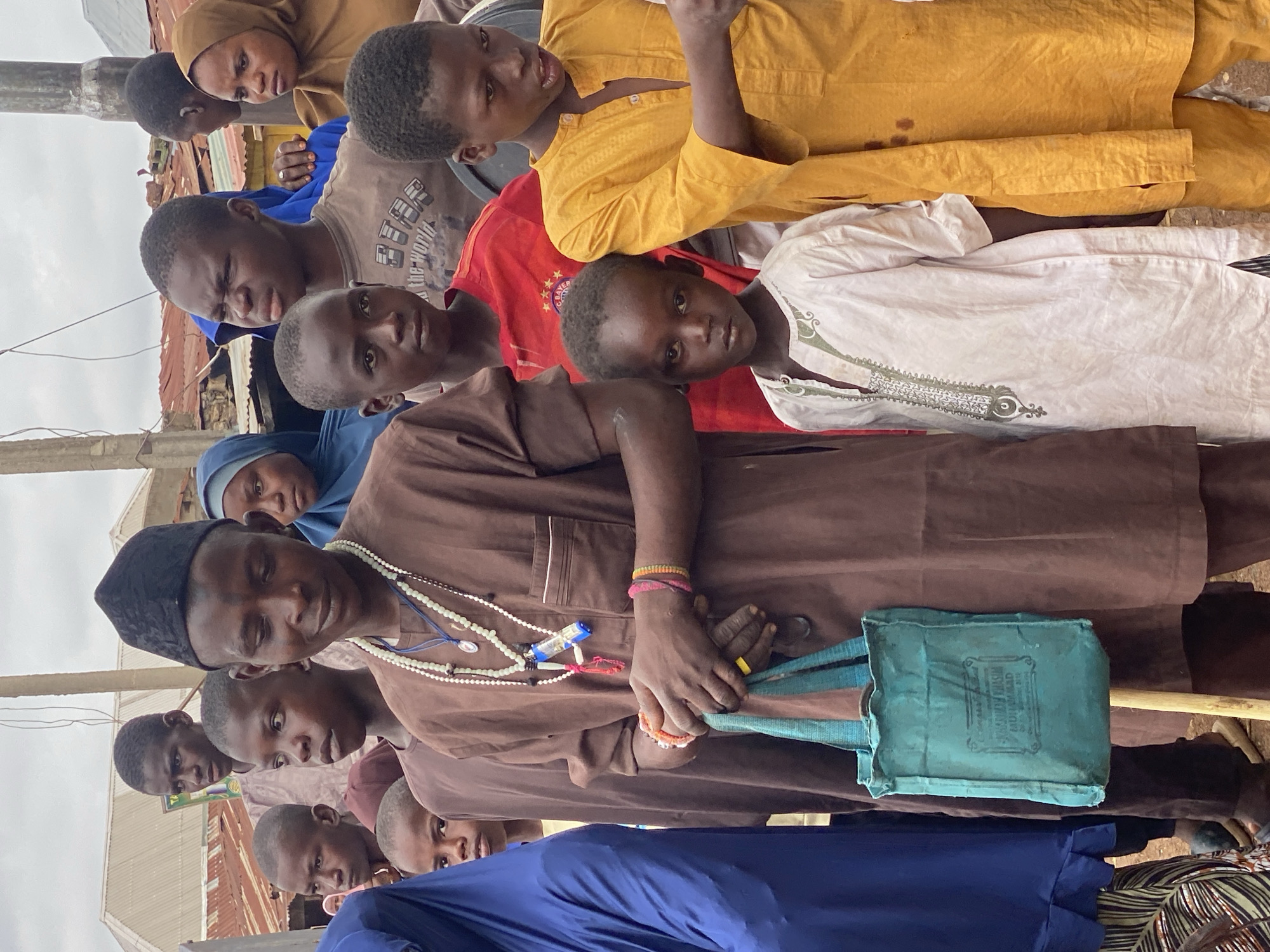 A group of people, including children and adults, stand together outdoors in what appears to be a market or community gathering in Rigachikun, Nigeria. The primary subject in the foreground is a man wearing a brown kaftan and a black cap, adorned with multiple strands of white beads and a small blue object around his neck. He is holding a light blue canvas bag with a stick. Several children are positioned around him, some looking towards the camera. To the right, a boy in a bright yellow kaftan stands prominently. The background reveals a collection of structures with corrugated metal roofs, characteristic of a semi-urban or rural Nigerian setting. The sky is overcast. The people are dressed in traditional clothing, with some wearing hijabs. The overall scene suggests a moment of pause or observation within a bustling community environment.