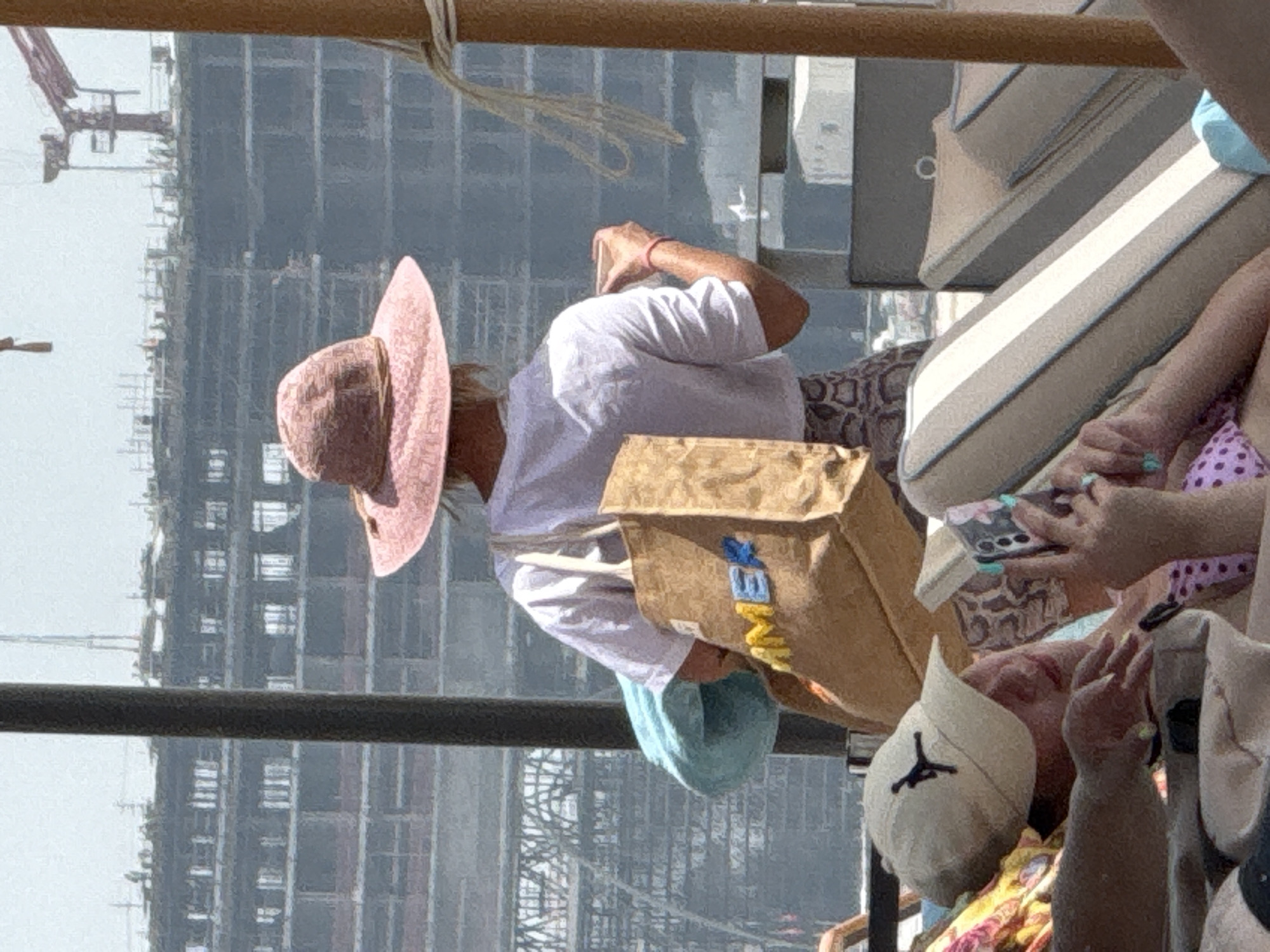 In an outdoor urban setting, a person wearing a wide-brimmed pink hat and a white t-shirt stands with their back to the camera, holding a brown tote bag with colorful letters spelling "SUMMER" and a light blue towel. They are looking towards a large construction site with multiple floors and cranes visible. In the foreground, a person wearing a tan baseball cap with a black logo and a woman in a bikini are relaxing on lounge chairs. The woman is holding a smartphone and appears to be taking a photo. The environment suggests a sunny day at a beach or pool area within the United Arab Emirates, possibly in a developing district of Dubai, given the extensive construction.