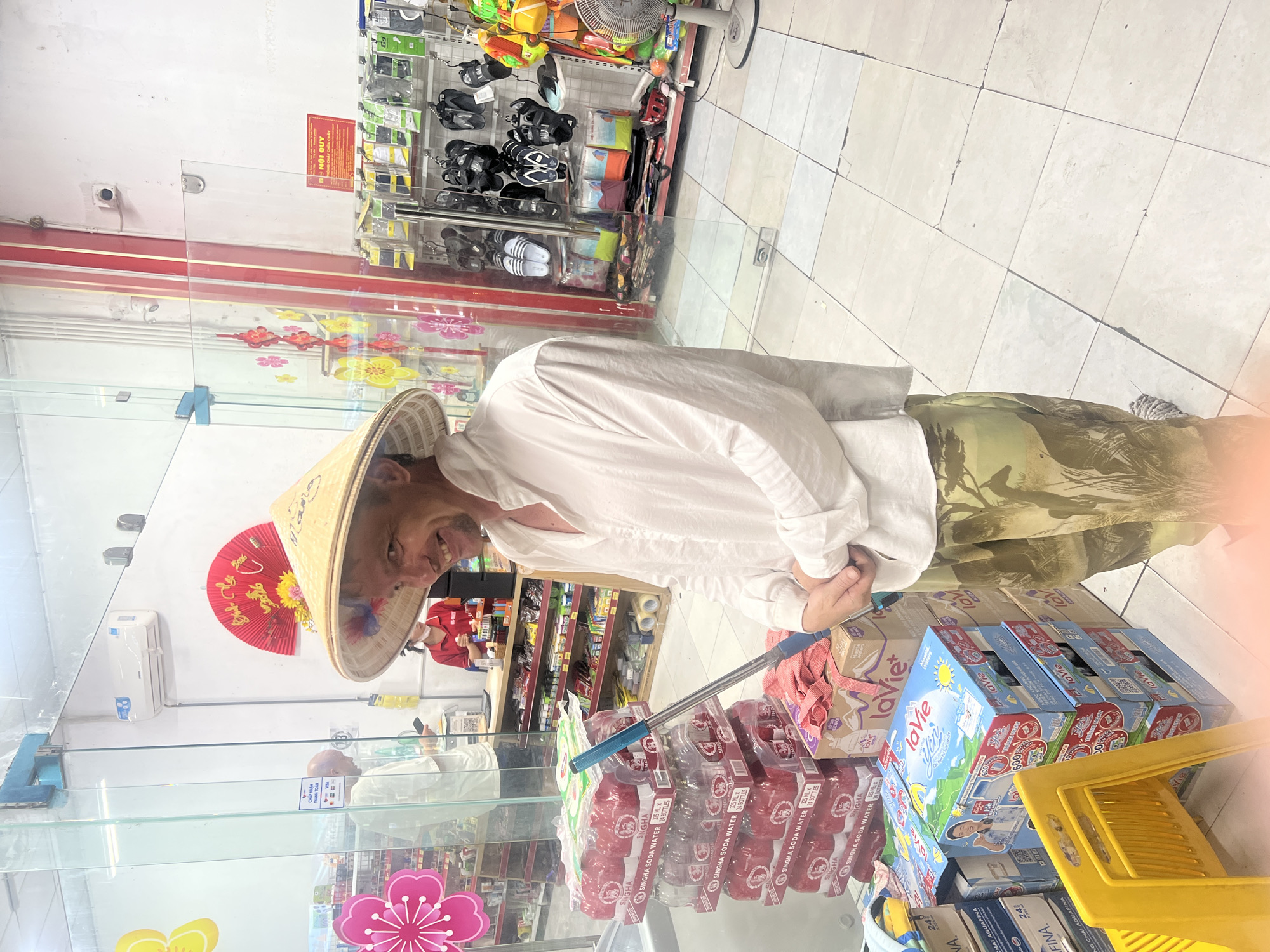 A smiling man wearing a conical hat and a white shirt stands in a convenience store in Da Nang, Vietnam. He is wearing camouflage pants and holding a mop. In the background, shelves are stocked with various items, including drinks and shoes. A cashier is visible behind a counter, and decorations for a holiday, possibly Tet, are displayed. The store has a tiled floor and glass doors.