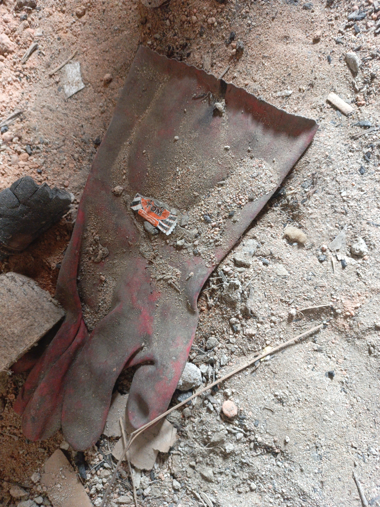 The image presents a top-down view of a construction site or demolition area in Unknown, Unknown. The main subject is a large, soiled red industrial glove lying on a mixture of sand, gravel, and debris. Adjacent to the glove are fragments of charcoal, wood, and brick. A wrapper from a TOM brand candy lies on the glove. The environment suggests a neglected or abandoned space undergoing renovation or demolition.