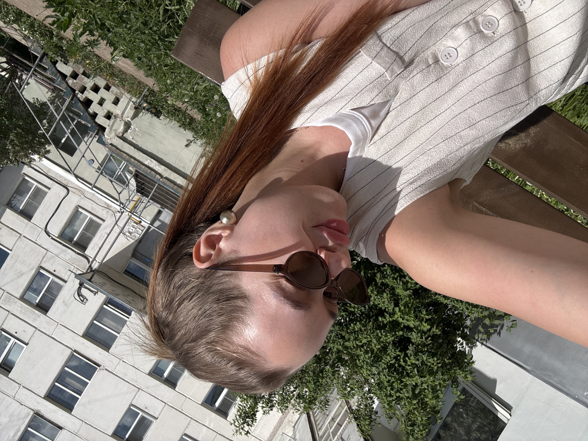 A fair-skinned woman is sitting on a wooden bench outdoors. She has light brown hair with auburn highlights and is wearing round sunglasses with a brown frame. She also has a large pearl earring. She is wearing a striped shirt or dress that has buttons. In the background, there are buildings and green trees. The photo was taken in Chaikovsky, Russia.