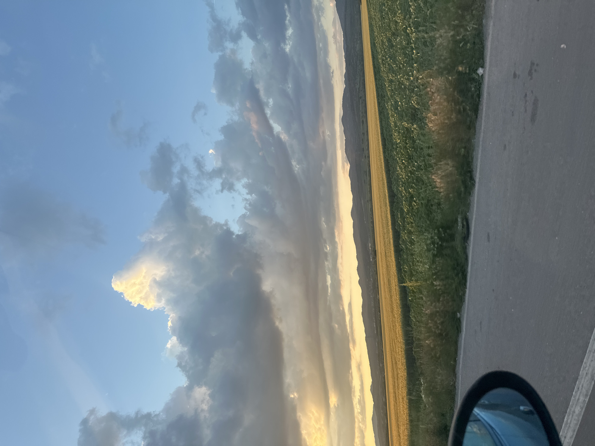 The image captures a rural landscape near Lokorsko, Bulgaria, observed from a vehicle during late afternoon or sunset. The upper two-thirds of the frame are dominated by a sky featuring scattered cumulus and stratus clouds. On the left, prominent cumulus clouds are brightly illuminated with golden-yellow hues by the low sun, contrasting with darker grey clouds extending across the remainder of the sky. A faint crescent object, possibly the moon, is visible in the upper right portion of the blue sky.
A line of dark, undulating hills or low mountains forms the horizon line across the middle distance. Below these mountains, two distinct agricultural fields stretch horizontally. The field on the left and center is a golden-yellow, indicative of mature grain, intensely lit by the setting sun. To the right of this, a second field exhibits a vibrant green color with visible rows of plants, possibly young sunflowers, appearing partially shadowed.
The foreground features an asphalt road, dark grey in color, with a faint white line along its left edge. A strip of uncultivated vegetation, consisting of various green and dried grasses and weeds, separates the road from the fields. A small, crumpled white object is discernible on the roadside near the green field. In the lower left corner, the black frame and reflective surface of a vehicle's side mirror are visible, indicating the point of view. No individuals are present in the scene.