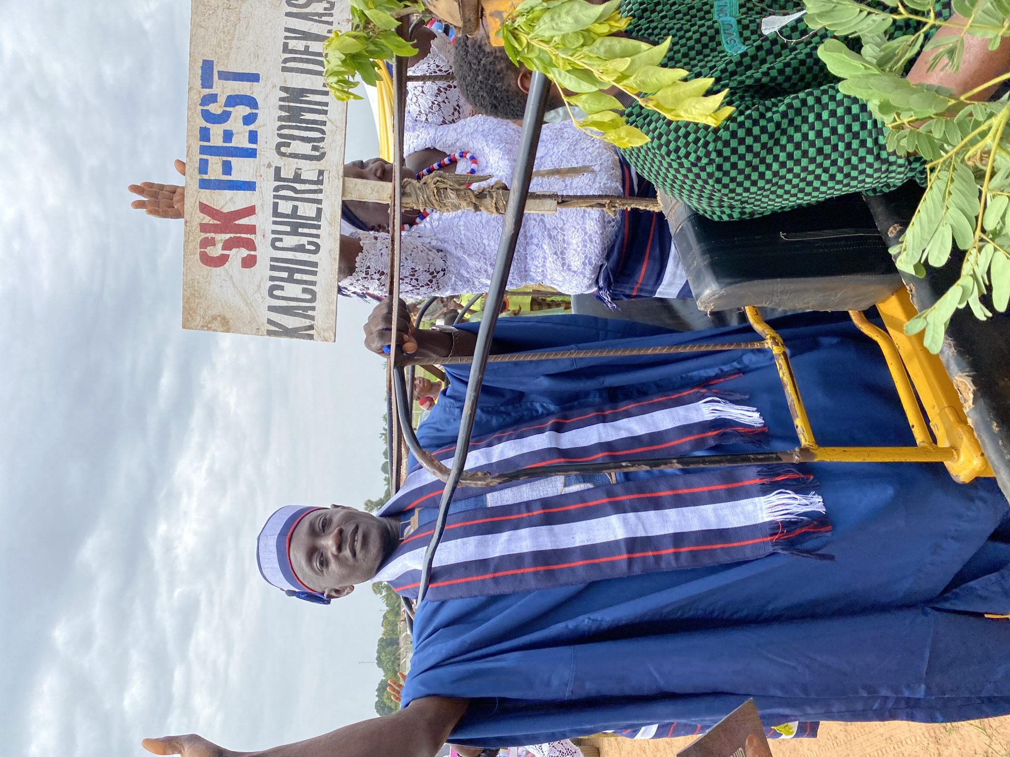 In Kafanchan B, Nigeria, the image shows a man wearing a blue robe and matching cap, likely traditional attire. He stands next to a yellow, possibly makeshift, platform. He appears to be looking toward the camera, and his hand is raised slightly, as if in greeting. Behind him, a sign reads "SK-FEST" in large, colorful letters, with "KACHICHERE COMM. DEVAS" underneath in smaller, plainer font. Another person is partially visible behind the man, wearing white lace clothing and a beaded necklace. Green foliage partially obscures the view. The background shows a cloudy sky and indistinct trees in the distance.