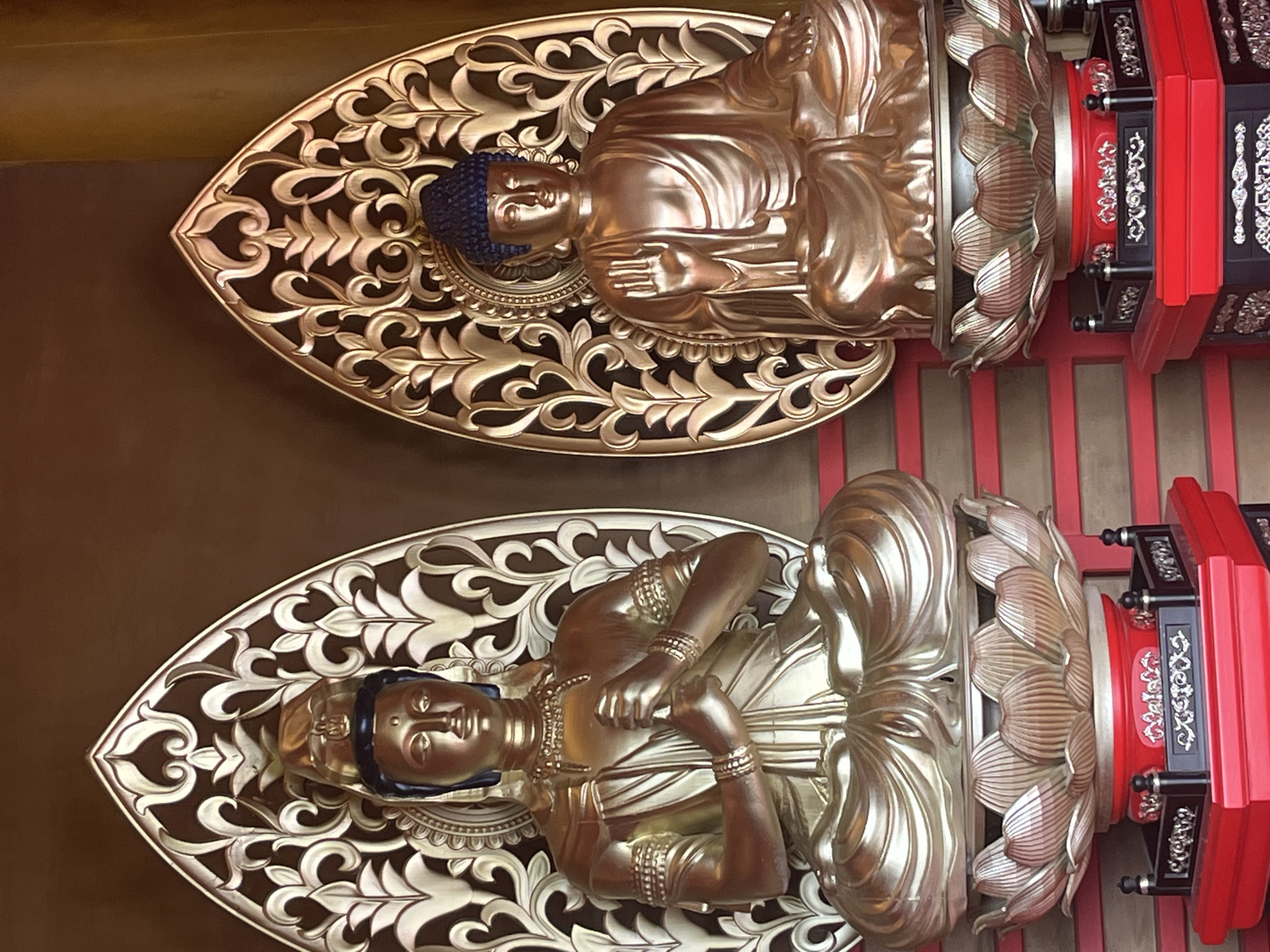 The image displays two golden Buddhist statues seated on lotus flower pedestals. The statue on the left has its hands clasped in front of its chest, while the statue on the right has its right hand raised with fingers extended upwards. Both statues are adorned with intricate jewelry and wear monastic robes. They are positioned against a dark brown background, with ornate golden halos behind them featuring decorative scrollwork. The pedestals are red and black, with decorative silver motifs. The statues appear to be indoors, possibly in a temple or religious establishment. There is no indication of the city of Krasnodar, Russia, in the image or its surrounding context.