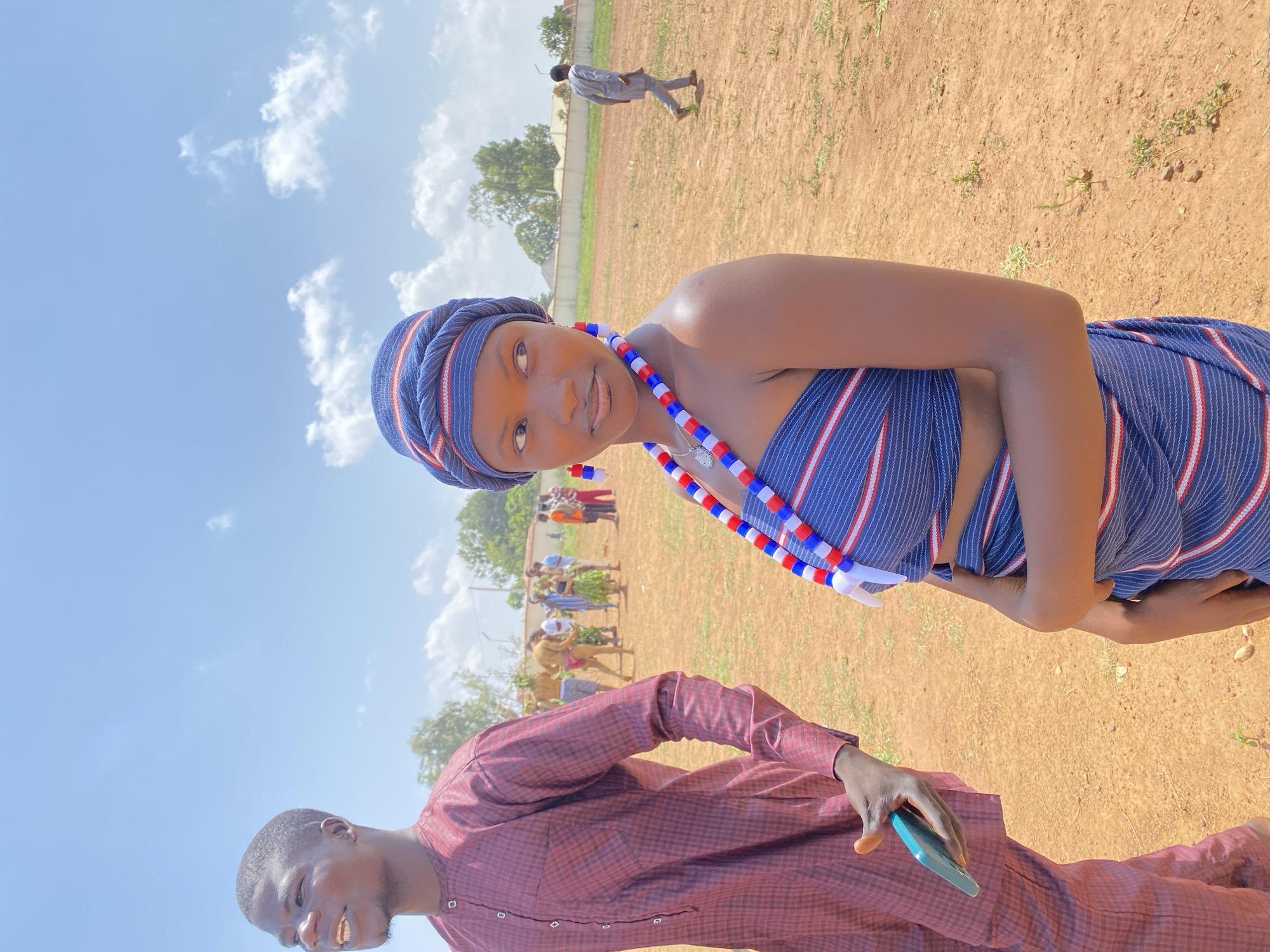 The image shows two people in an outdoor setting in Durumi, Nigeria, with a reddish-brown ground and scattered green vegetation. In the foreground, a man wearing a maroon, button-down shirt is visible from the chest down; he is holding a blue mobile phone. Adjacent to him and slightly above, a woman wearing a blue and white striped top and matching headwrap with a red, white, and blue beaded necklace is looking at the camera. In the background, a small group of people appear to be gathered, some holding objects. Another individual is walking separately further away. The sky is blue with scattered white clouds.
