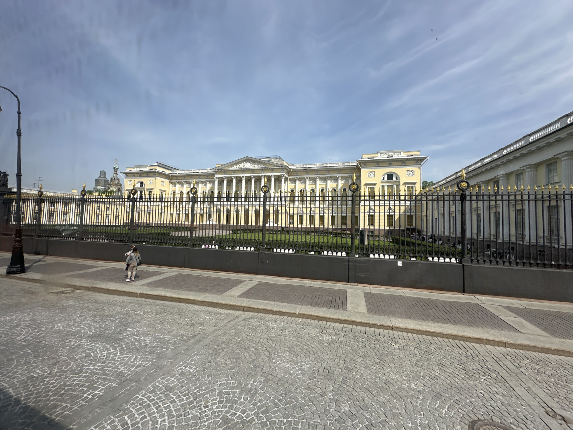 The image depicts a street scene in Saint Petersburg, Russia, featuring the facade of a grand, yellow building with white columns, likely a palace or government structure. A tall, ornate black iron fence runs along the perimeter of the building, separated from the cobblestone street by a sidewalk with interspersed paving stone.
A person stands on the sidewalk, facing the building and appears to be wearing light colored attire.
The sky is partly cloudy, suggesting a sunny day. In the background, adjacent to the primary building, are glimpses of other structures also with decorative exteriors. A lamppost stands near the edge of the frame on the left side.