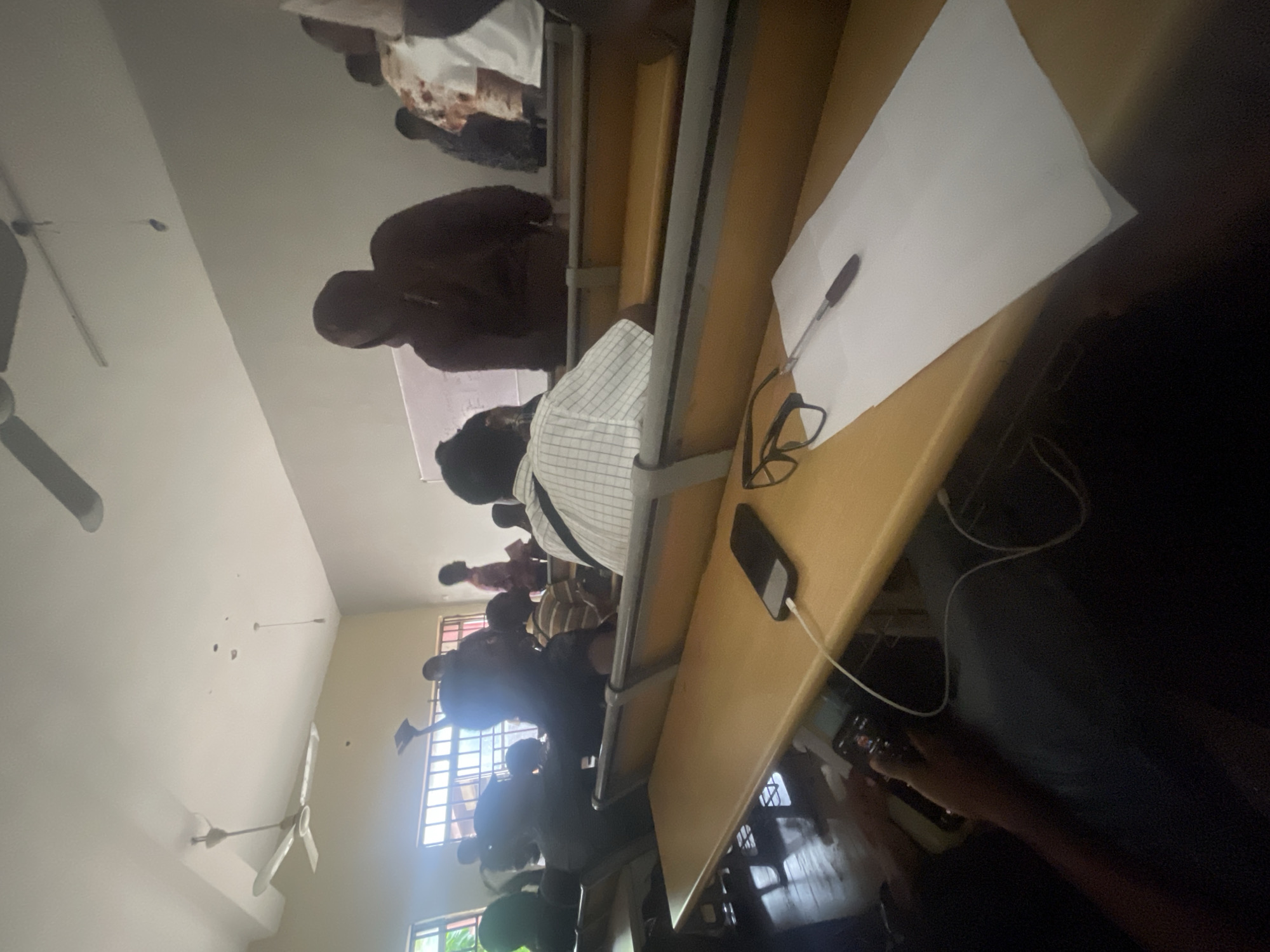 A medium shot captures an indoor scene, likely a lecture hall in Ihiagwa, Nigeria, with rows of wooden desks and benches. Several individuals are seated, facing forward, and some are standing. The room has a high ceiling with white paint, and a ceiling fan is visible. Natural light streams in from windows with barred frames.
In the foreground, a wooden desk holds a black smartphone connected to a charger, a pair of black-rimmed glasses, a pen, and a sheet of white paper. To the left, a person's hands are visible holding another smartphone. The background reveals more students, some of whom appear to be looking at a whiteboard. The overall atmosphere suggests a typical academic setting during a lecture or class session.