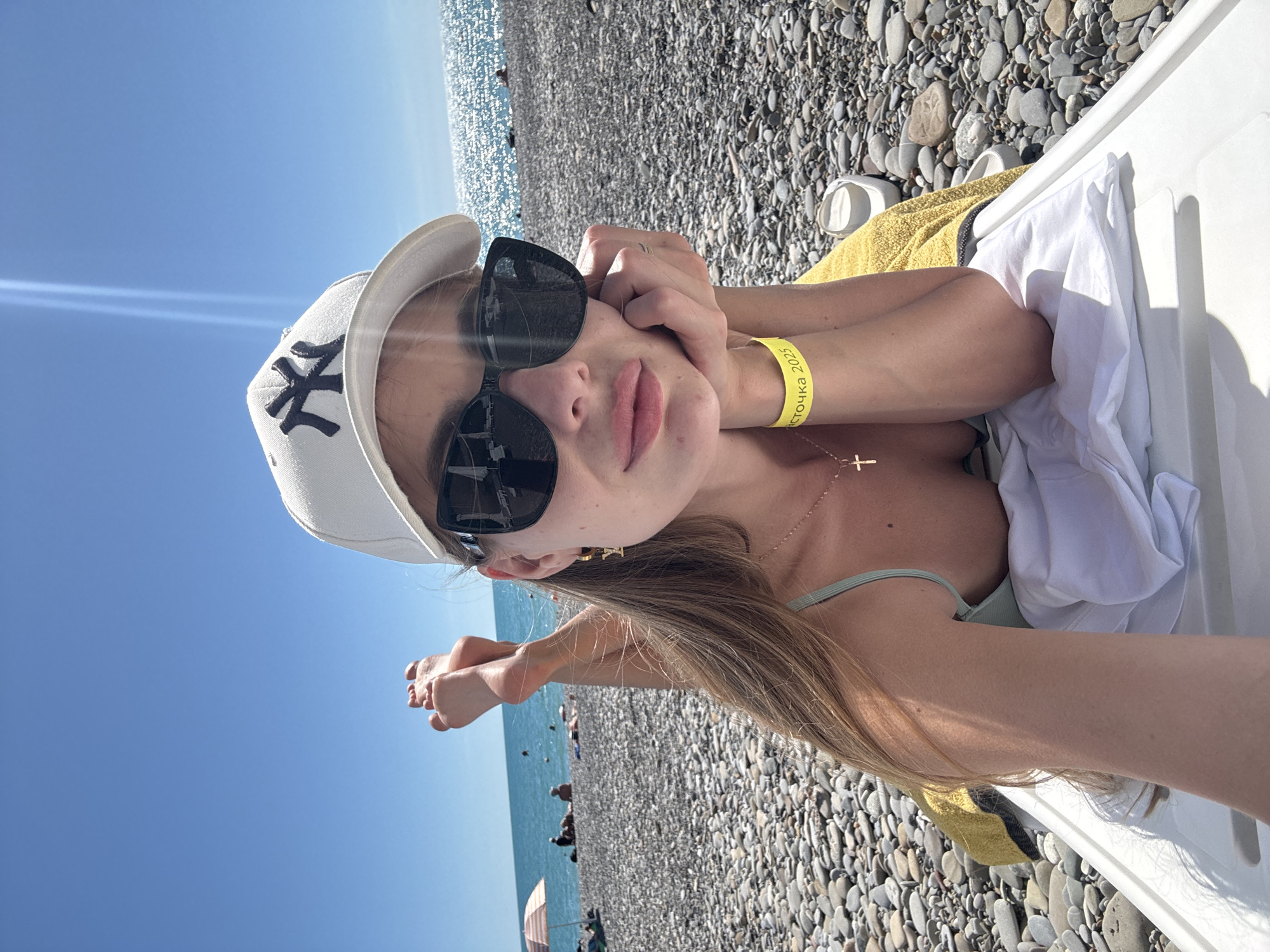 A young woman with blonde hair, wearing a white baseball cap with a black logo, sunglasses, and a light green bikini top, is lying on a white lounge chair on a pebble beach. She is propped up on her elbow, with her hand resting on her chin, and her feet are raised in the air. A yellow towel is underneath her. She is wearing a gold necklace with a cross pendant and a yellow wristband. In the background, the blue sea and a clear sky are visible, with a few people and beach umbrellas in the distance. The scene is set on a beach in Sochi, Russia, indicated by the wristband that reads "восточка 2025", which translates to "Vostochny 2025".