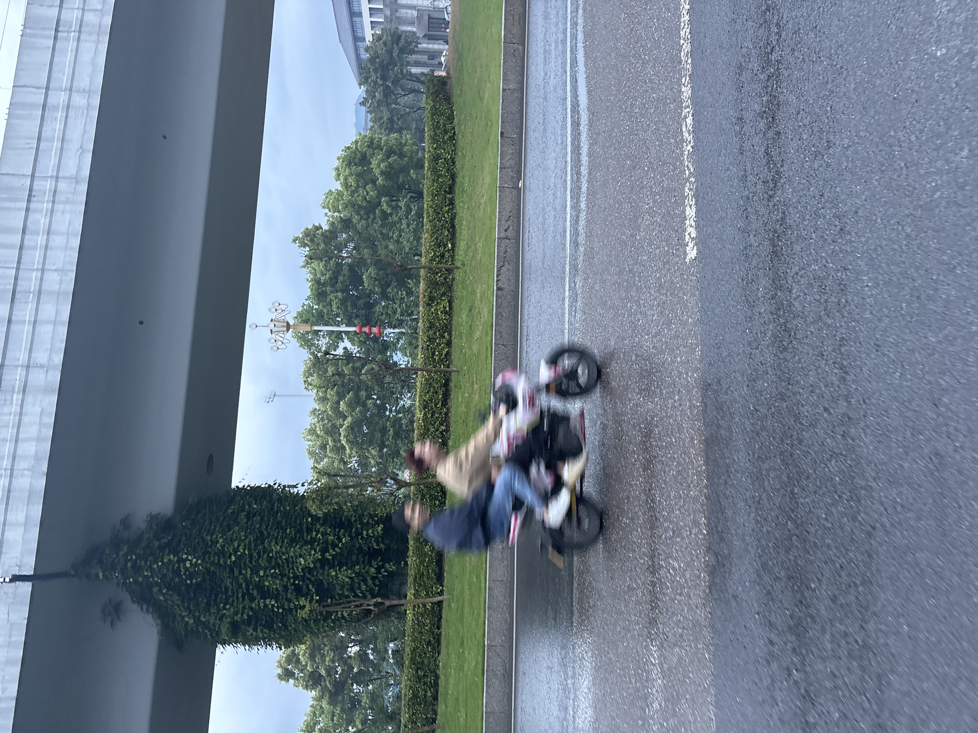 A motion-blurred photograph depicts two individuals riding a tricycle on an asphalt road in Jiangjin, China. The road features a white line marking. Green foliage borders the roadway, with trees visible in the background. Above, a concrete overpass partially obscures the sky and reflects a distorted image of the foliage below. The scene appears to be captured during or after rainfall, as the road surface is wet. The speed of the camera is not capturing any details and it is more difficult to describe the people and tricycle.