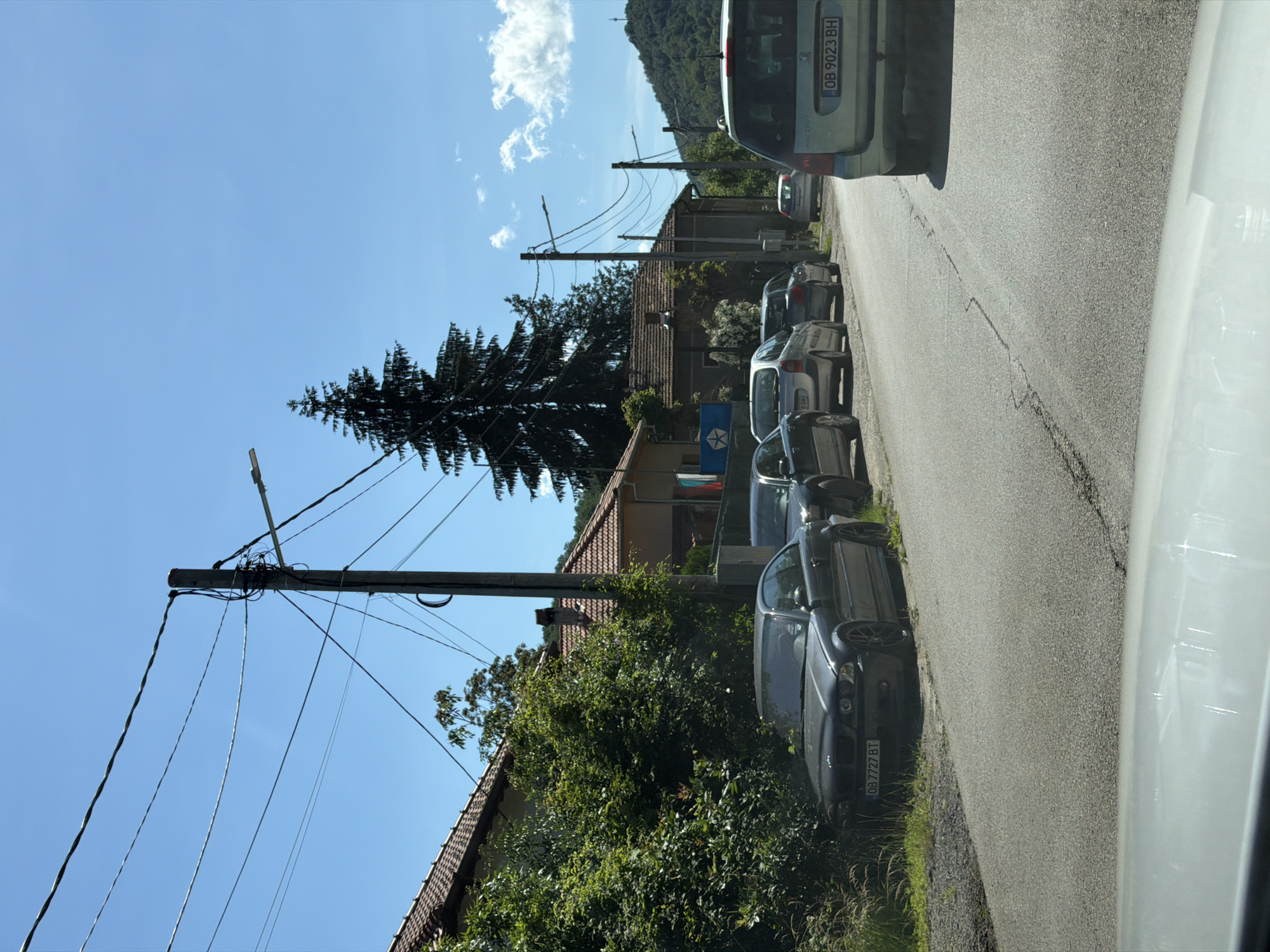 In Beli Osam, Bulgaria, a street scene unfolds under a clear blue sky. Several parked cars line the paved road, which has visible cracks and wear. The vehicles include a dark gray BMW coupe with license plate "OB 7727 BT" parked on the left, followed by a silver hatchback, another silver car, and a light green minivan with license plate "OB 9023 BH" further down the road.
On the left side of the street, houses with red-tiled roofs are visible, partially obscured by lush green trees and bushes. A large, dark green evergreen tree stands tall in the background. Utility poles with numerous wires crisscross the sky, and streetlights are mounted on some of them. In the distance, rolling green hills form the backdrop. Two flags are visible hanging from one of the houses, one appears to be the Bulgarian flag. The scene appears to be a quiet residential area.