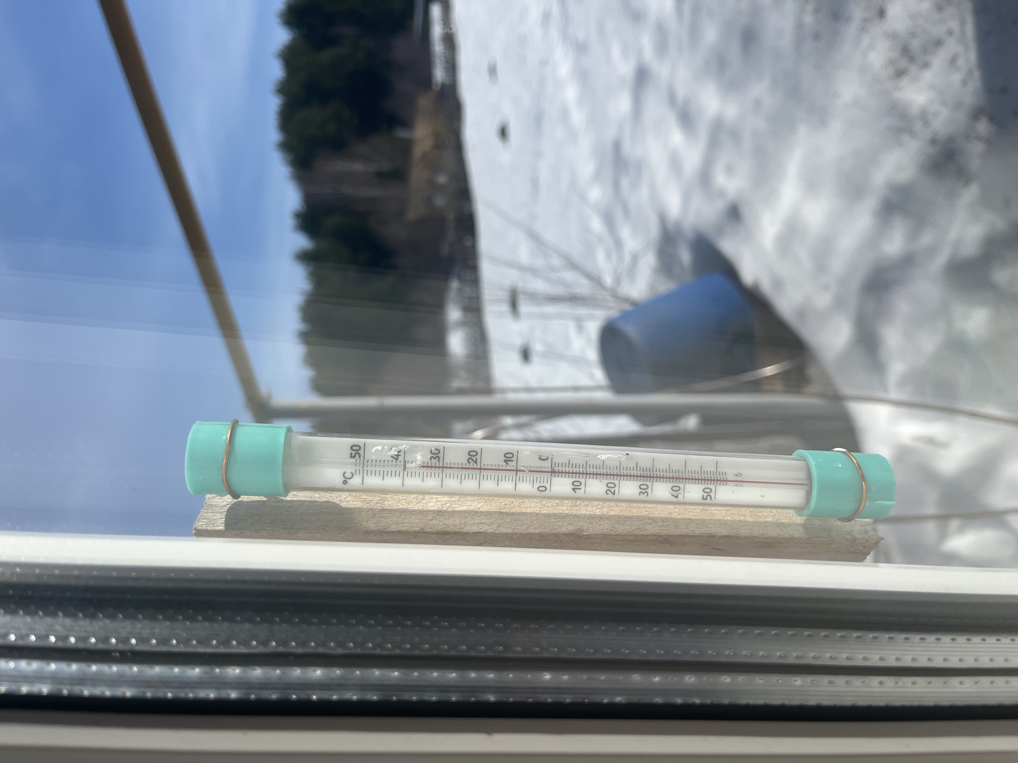 A photograph taken indoors shows a glass thermometer placed on a small wooden block sitting on a windowsill. The thermometer reads approximately 6 degrees Celsius. Outside the window, there is a snow-covered landscape with trees and a blue object, possibly a bucket, is visible. The location is Большой Букор, Russia.