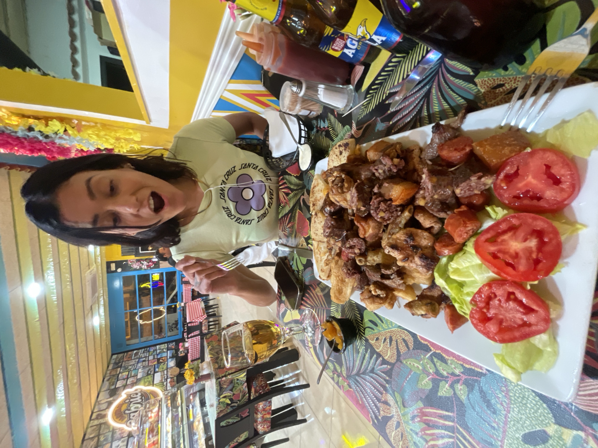 A woman is about to eat a meal consisting of a variety of fried meats and vegetables on a white plate. Sliced tomatoes and lettuce are also visible. The woman is holding a fork and knife and is facing the camera. Behind her are other tables and chairs, and in the distance, a window and an illuminated sign with the name "Gabriel." Several bottles and condiments are on the table near the food plate. The restaurant is located in L'Hospitalet De Llobregat, Spain. The table has a colorful patterned tablecloth.