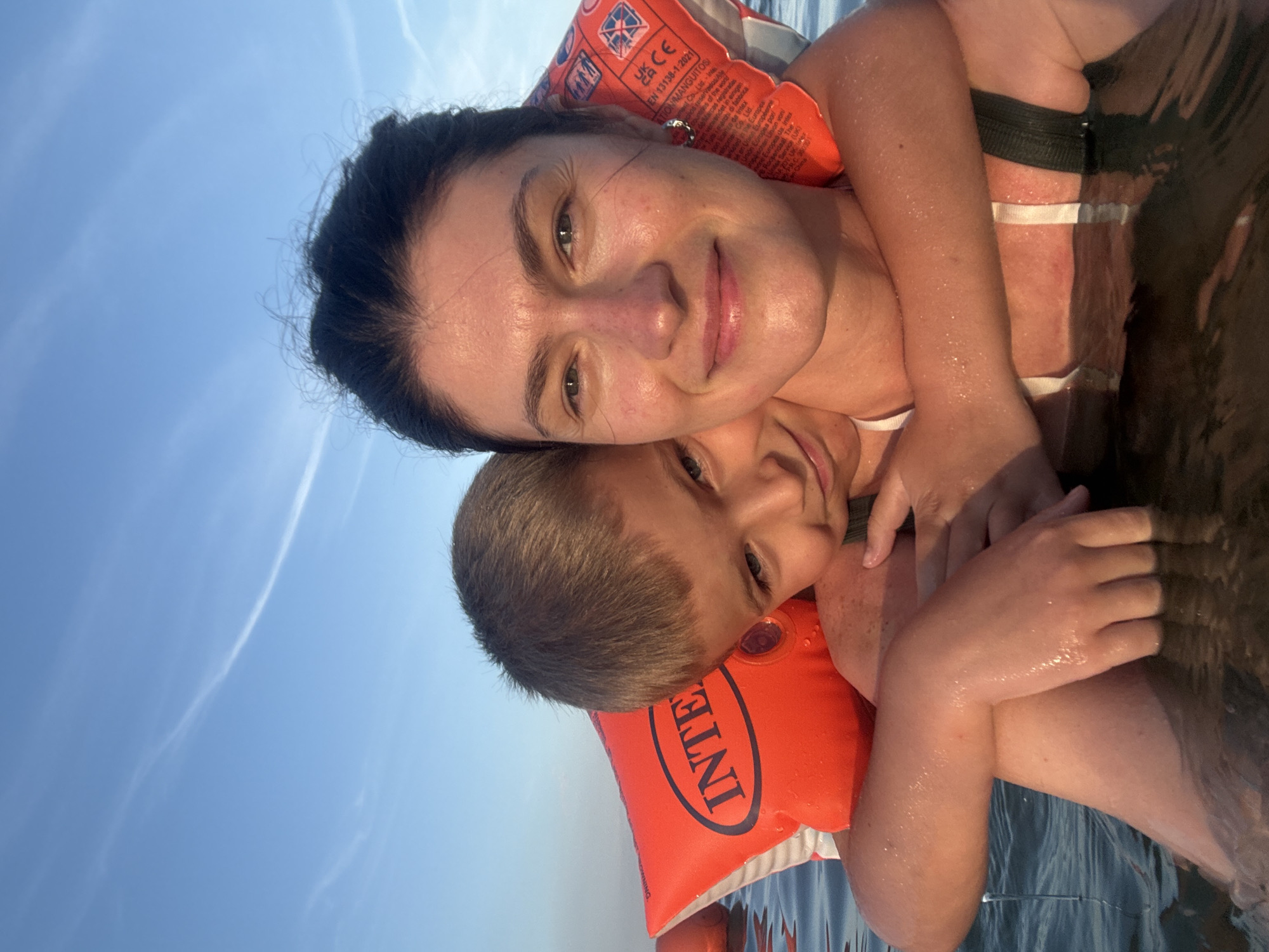 A selfie captures a smiling woman and a young boy in the water, likely Nha Trang, Vietnam, given the context. The woman, with dark hair and fair skin, is looking directly at the camera, her face illuminated by natural light. She wears a dark swimsuit. The boy, positioned behind her, has blonde hair and is hugging her from behind, his face also turned towards the camera. He is wearing orange arm floaties with the brand name "INTE." The water is calm, with subtle ripples and reflections. The sky above is clear blue with a few wispy white clouds. The overall impression is one of a happy, relaxed moment shared between the two individuals.