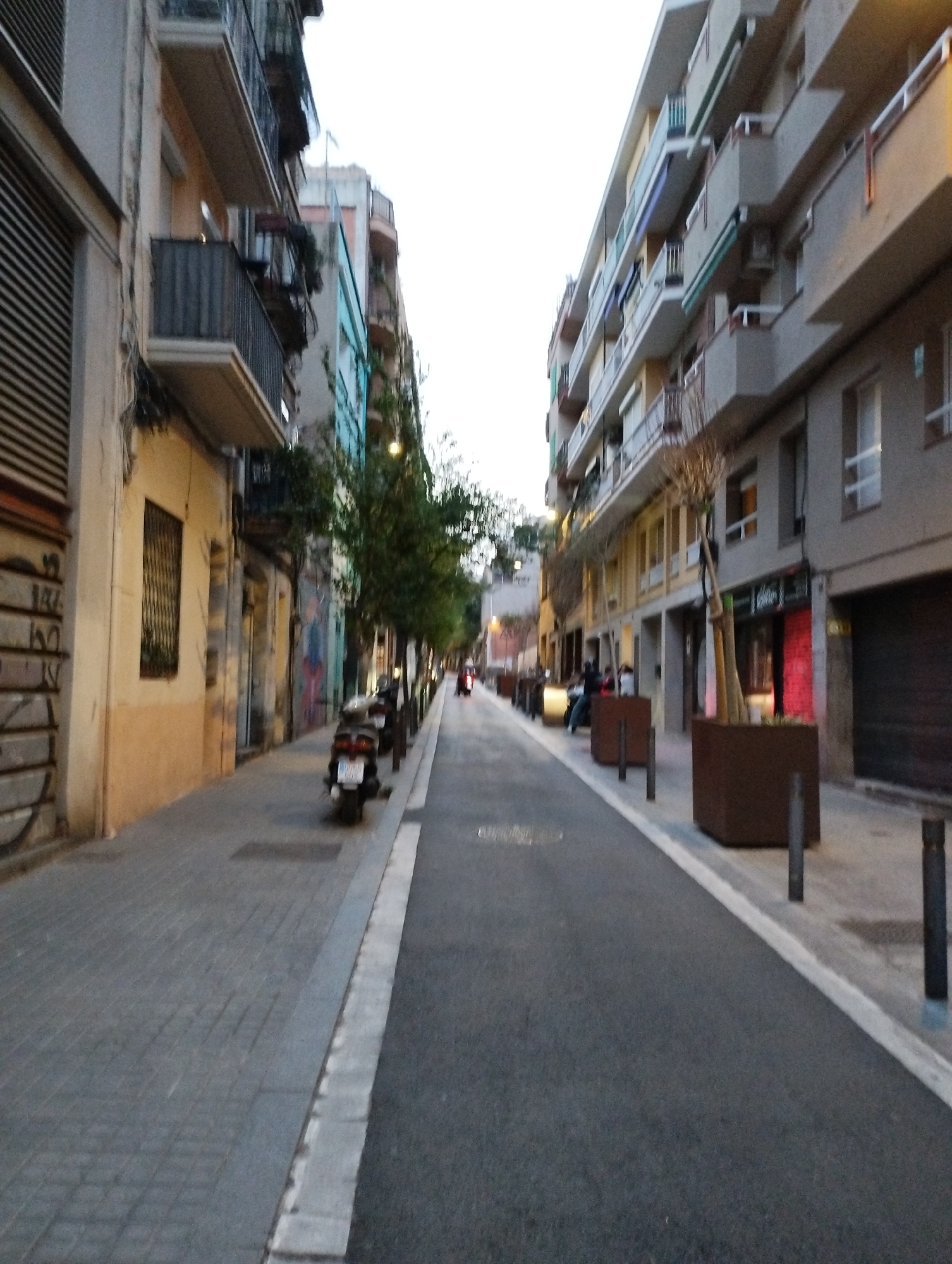 The image shows a street scene in Barcelona, Spain, likely in the Eixample district or a similar residential area with a mix of older and newer apartment buildings. The perspective is from the street level, looking down a relatively narrow street lined with buildings on both sides.
On the left, a building features a beige facade with horizontal blinds and a prominent balcony with dark metal railings. Next to it is a narrower building with a lighter facade, also with balconies. Further down this side of the street, there's a building with a pale yellow facade, a barred window, and signs of graffiti. The sidewalk on this side is paved with gray interlocking bricks.
On the right, a multi-story apartment building dominates the view. It has a series of balconies with concrete railings and some with metal railings, some featuring awnings. The ground floor of this building appears to house shops, with one visible entrance that is brightly lit with red light. Planter boxes with sparse trees are situated along the edge of the sidewalk on this side.
The street itself is asphalt with a white line marking the edge of the sidewalk. Several parked scooters are visible on the left sidewalk, one in the foreground. A few people are present in the background on the right side, near the shops, and one person is riding a scooter in the distance on the right. The sky is overcast, indicating it is likely early morning or late afternoon. The overall impression is of a residential urban street with commercial activity on the ground floor.
