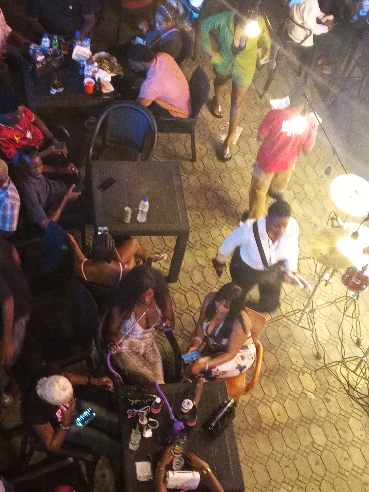 The image captures an aerial view of a gathering at night, possibly an outdoor bar or restaurant, in Lokoja, Nigeria. A diverse group of individuals are seen seated and standing around tables, engaged in various activities. In the foreground, two women are seated, one of whom is holding a hookah pipe. Another woman with blonde hair is looking at her phone. Several other people are visible at different tables, some holding drinks and engaging in conversations. The environment appears to be paved with interlocking bricks, with string lights overhead illuminating the scene. The overall atmosphere suggests a social event or a night out.