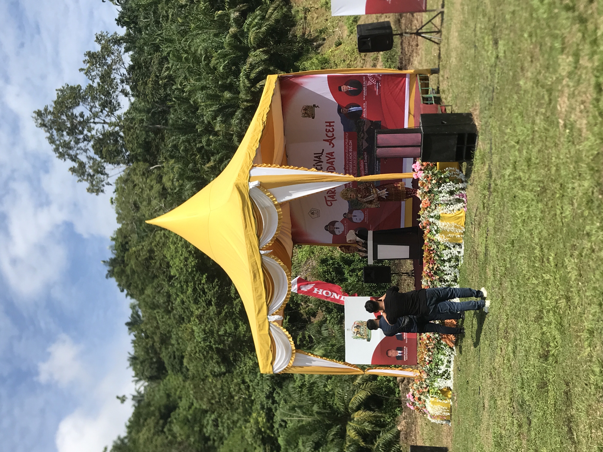 An outdoor event is taking place under a yellow canopy tent, set against a backdrop of lush green hills and trees. The tent has white drapes and is decorated with flowers. On a stage within the tent, a woman in traditional attire stands at a podium, speaking to an audience. Behind her, a banner reads "Festival Budaya Aceh" (Aceh Cultural Festival). Two men are standing in front of the stage, one appearing to take a photograph with his phone, while another man is partially obscured. Several black speakers are placed on the ground around the stage. The event appears to be held in a grassy area, likely a park or open field in Langkuta, Indonesia, given the cultural context of the banner. The sky is partly cloudy with a bright blue hue.