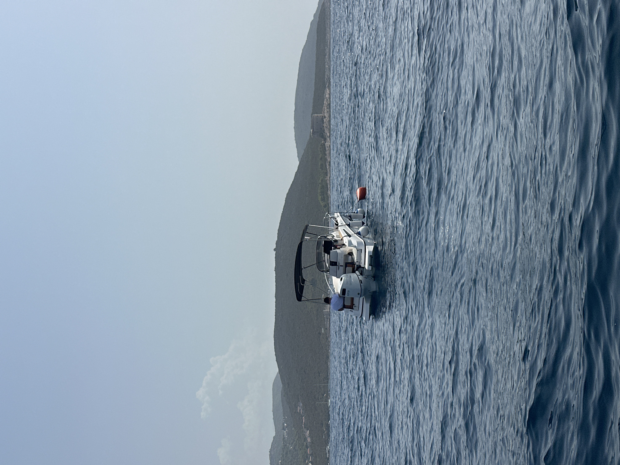 A white motorboat with a black canopy is anchored in calm blue water. Two individuals are seated on the boat. In the background, a hilly coastline with sparse vegetation and some buildings is visible under a hazy sky. A round orange buoy floats near the stern of the boat. The scene appears to be along the coast of Luštica, Montenegro, with a faint structure resembling a fort on a distant hill.