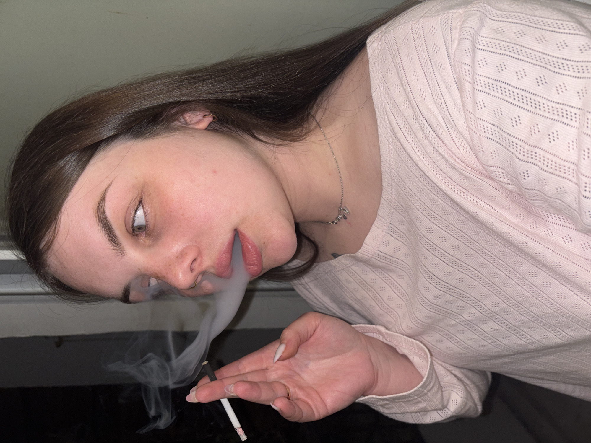 An indoor, close-up, and vertically oriented photo captures a young woman smoking a cigarette. The woman, with long, dark brown hair, fair skin, and light-colored eyes, is seen from a slightly elevated perspective, looking upwards. Her face shows a neutral to slightly contemplative expression, with her mouth slightly open as she exhales a plume of thick, white smoke. She has a small, gold hoop piercing in her left nostril and a gold hoop earring in her left ear.
She is wearing a light pink, long-sleeved shirt with a delicate, openwork pattern featuring horizontal lines and small diamond shapes. A thin silver necklace with a small, reflective pendant hangs around her neck. Her left hand, with long, light-colored fingernails, holds the cigarette between her index and middle fingers. The cigarette has a white paper casing and a dark filter, and its tip glows faintly. The background is dark, suggesting a dimly lit indoor setting, possibly at night or in the evening. The provided context indicates the location is Chornomorskoe, Ukraine. There is no visible text in the image.