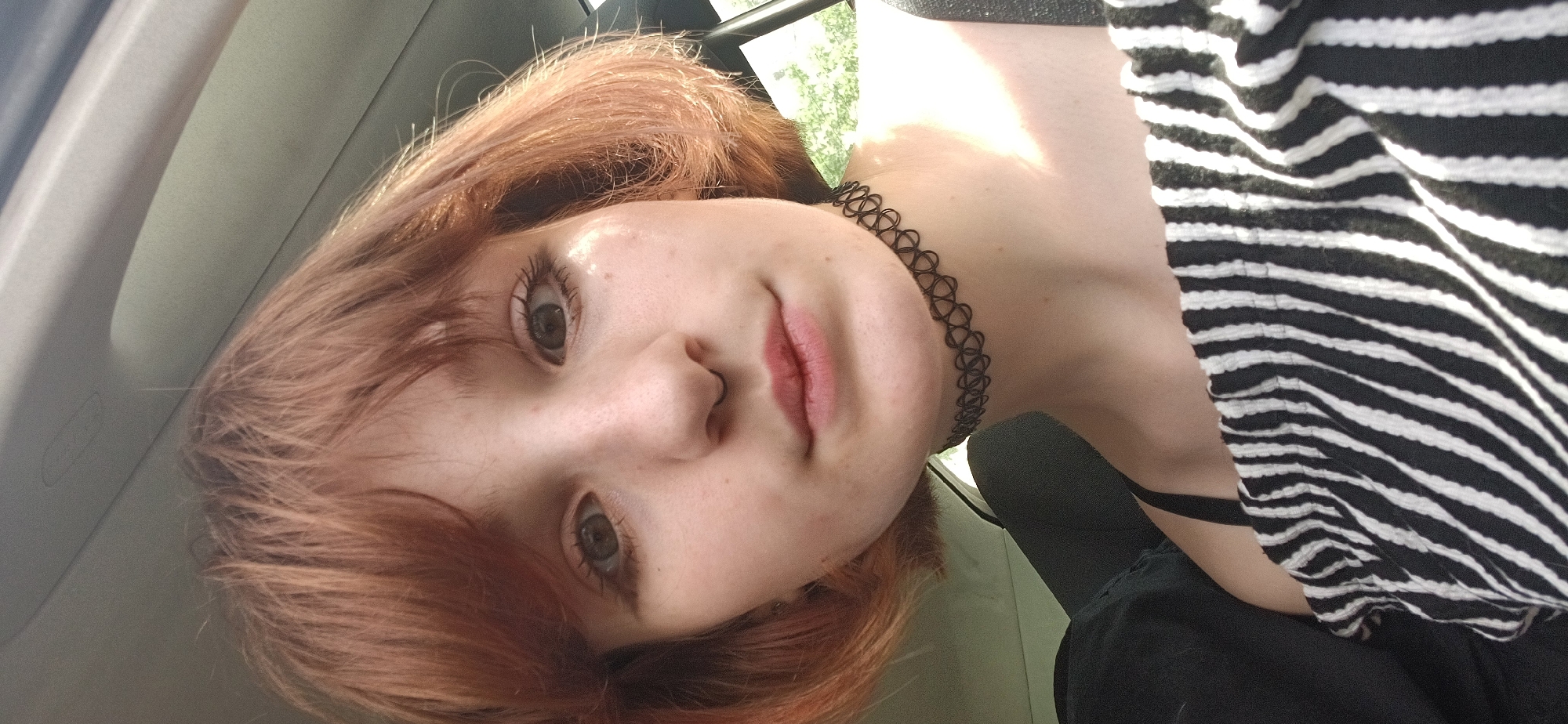 A young, fair-skinned person with short, reddish-brown hair is facing the camera. They are wearing a black choker necklace and a black and white striped top. The person is positioned inside a vehicle, as evidenced by the interior trim visible in the background. Outside the vehicle window, there is a glimpse of green foliage, suggesting a tree or other vegetation. The picture was not taken in the city of Bar, Montenegro.