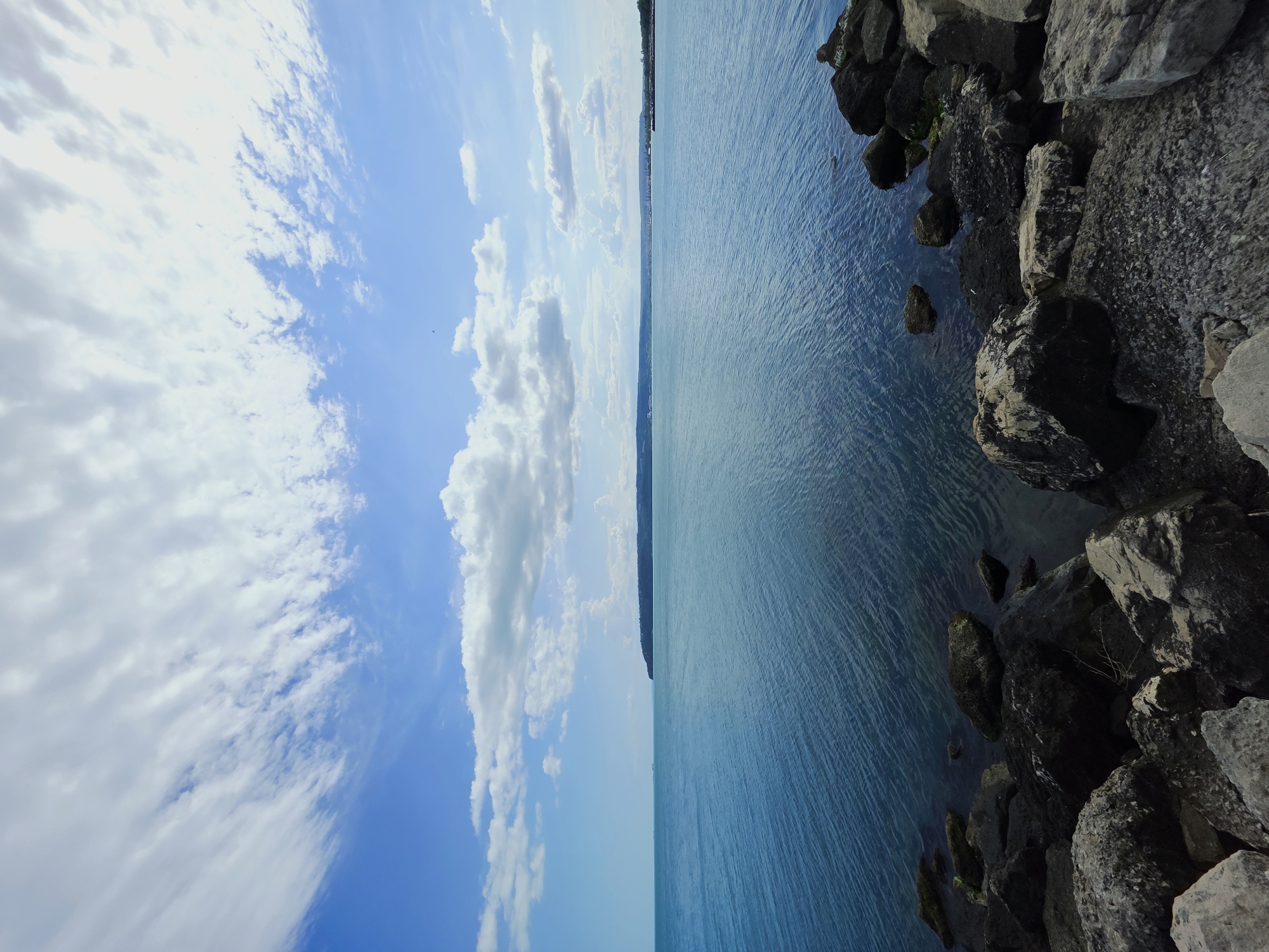 The image shows a scene in Varna, Bulgaria, specifically at a rocky shoreline. The foreground features large, rough rocks extending into the turquoise water of the Black Sea. The sea extends into the distance, meeting a distant shoreline. To the left, the sky is visible, filled with white clouds against a bright blue backdrop. The perspective suggests the photo was taken from a low angle, emphasizing the scale of the rocks and the expanse of the sky and sea. No people or animals are visible.