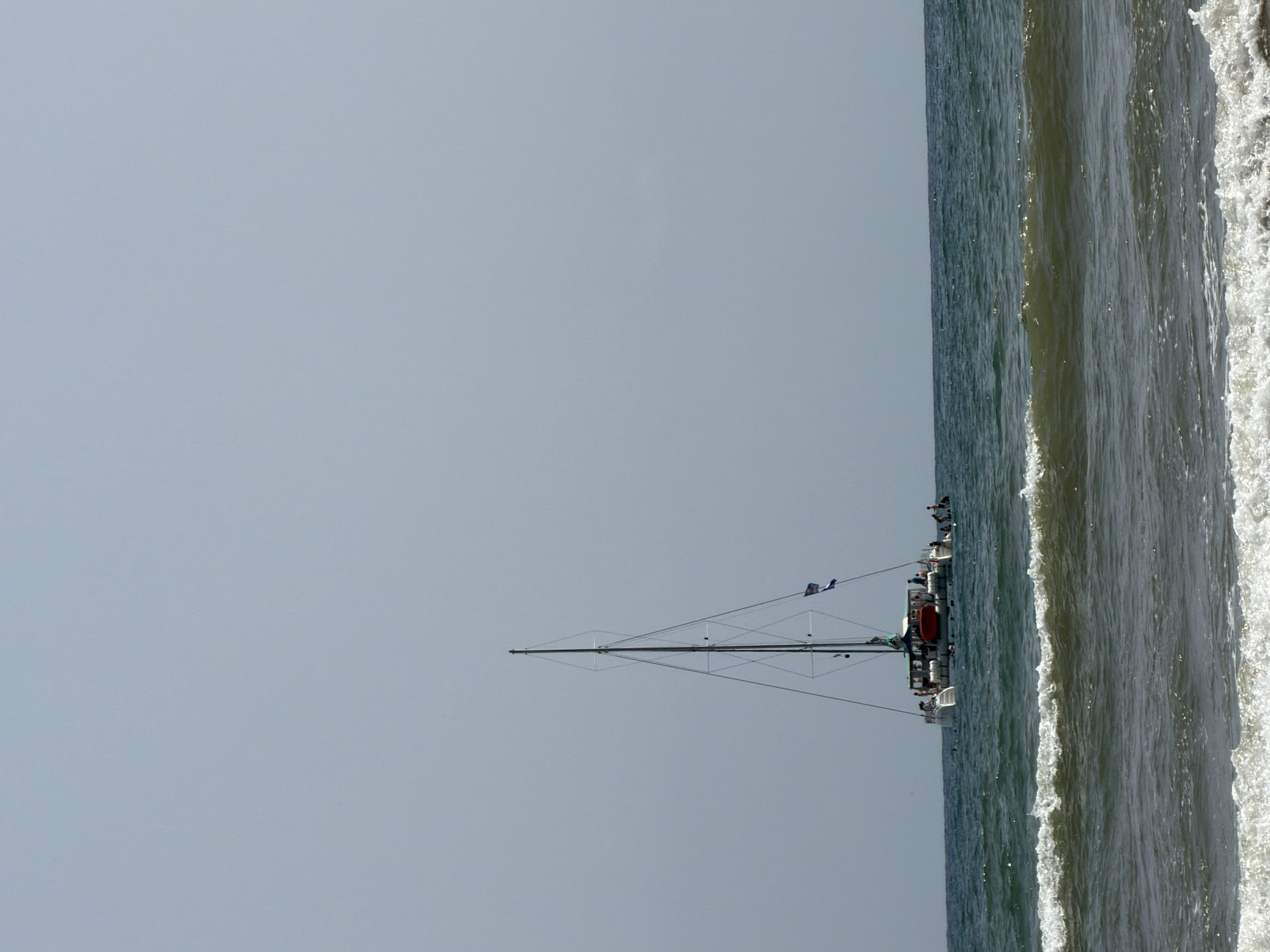 A wide shot shows a clear, overcast sky over a calm sea with moderate waves. In the middle ground, a white catamaran with multiple people aboard is sailing. The catamaran has a tall mast with two flags flying near the top. Several individuals are visible on the deck of the boat, some standing and others seated. The boat is positioned to the left of the frame. The foreground shows foamy white waves breaking. The overall scene depicts a daytime view of a boat on the ocean.
