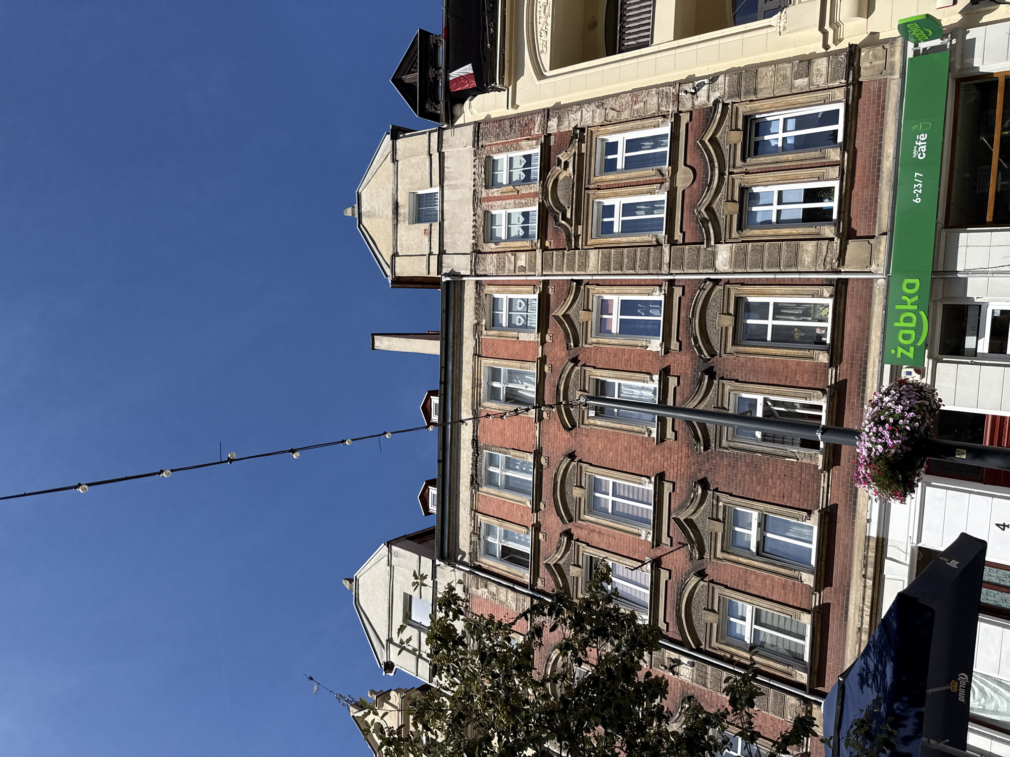 The image captures a street-level view of buildings on a sunny day in Zgorzelec, Poland. A string of decorative lights stretches across the bright blue sky. The facades of several buildings are visible, characterized by red brickwork and ornate architectural details around the windows. The windows themselves are predominantly white and appear to be in good condition.
On the ground floor, a green storefront with the "Żabka" logo is prominently displayed. This suggests a convenience store or a similar retail establishment. The text "6-23/7 żabka cafe" is also visible on the signage, indicating extended operating hours and a café service.
A large, dark blue umbrella, possibly part of an outdoor seating area for the cafe, is partially visible in the lower left of the frame. To the right of the umbrella, a black lamppost stands adorned with a hanging basket of colorful flowers. The street number "4" is visible on the building below the umbrella.
The buildings exhibit a mix of architectural styles, with some showing signs of age and repair, particularly around the upper floors where brickwork is exposed. The overall impression is of a typical European urban street scene, with a blend of historic architecture and modern commercial activity. No people are explicitly visible in the image.