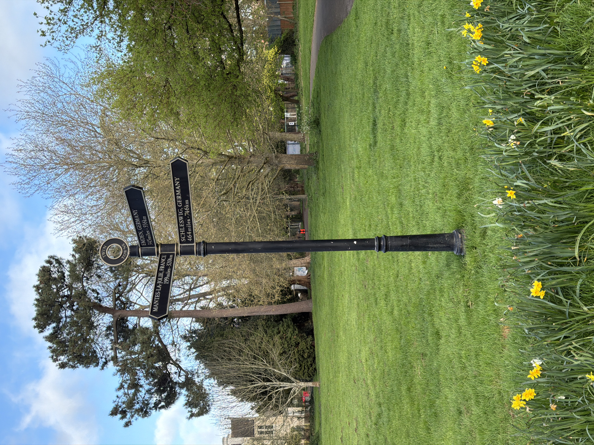This photograph depicts a black signpost situated in a grassy area, likely a park, with trees and daffodils in the background. The signpost has a circular emblem at the top with the inscription "TWIN TOWNS," followed by three directional signs indicating distances to various locations: Mantes-la-Jolie in France (199 miles/320 km), Emden in Germany (345 miles/556 km), and Schleswig in Germany (464 miles/746 km). The sky is partly cloudy, and the presence of daffodils suggests it is springtime. The photograph doesn't show the specific location in London, only the place name on the sign.