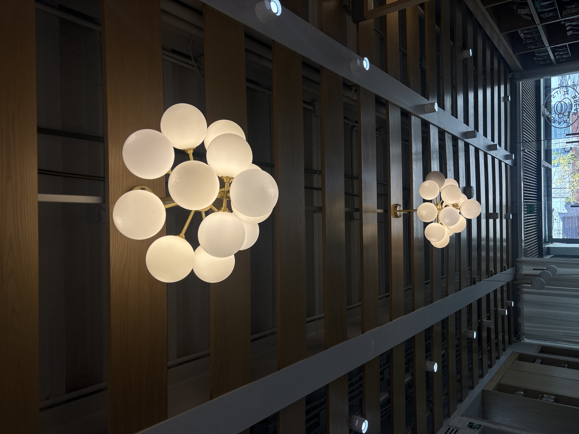 The image shows an interior space with a high ceiling, featuring a series of parallel wooden slats that create a textured and visually interesting effect. Hanging from the ceiling at different points are spherical light fixtures with multiple bulbs. The background is a mix of natural light from a window and architectural elements, including possible store shelves or displays. The image is taken from a low angle, looking upwards towards the ceiling. In the distance, a window reveals an urban environment, including a building with a sign that appears to be in Spanish. Based on this sign, the location is likely in Mexico City, Mexico.