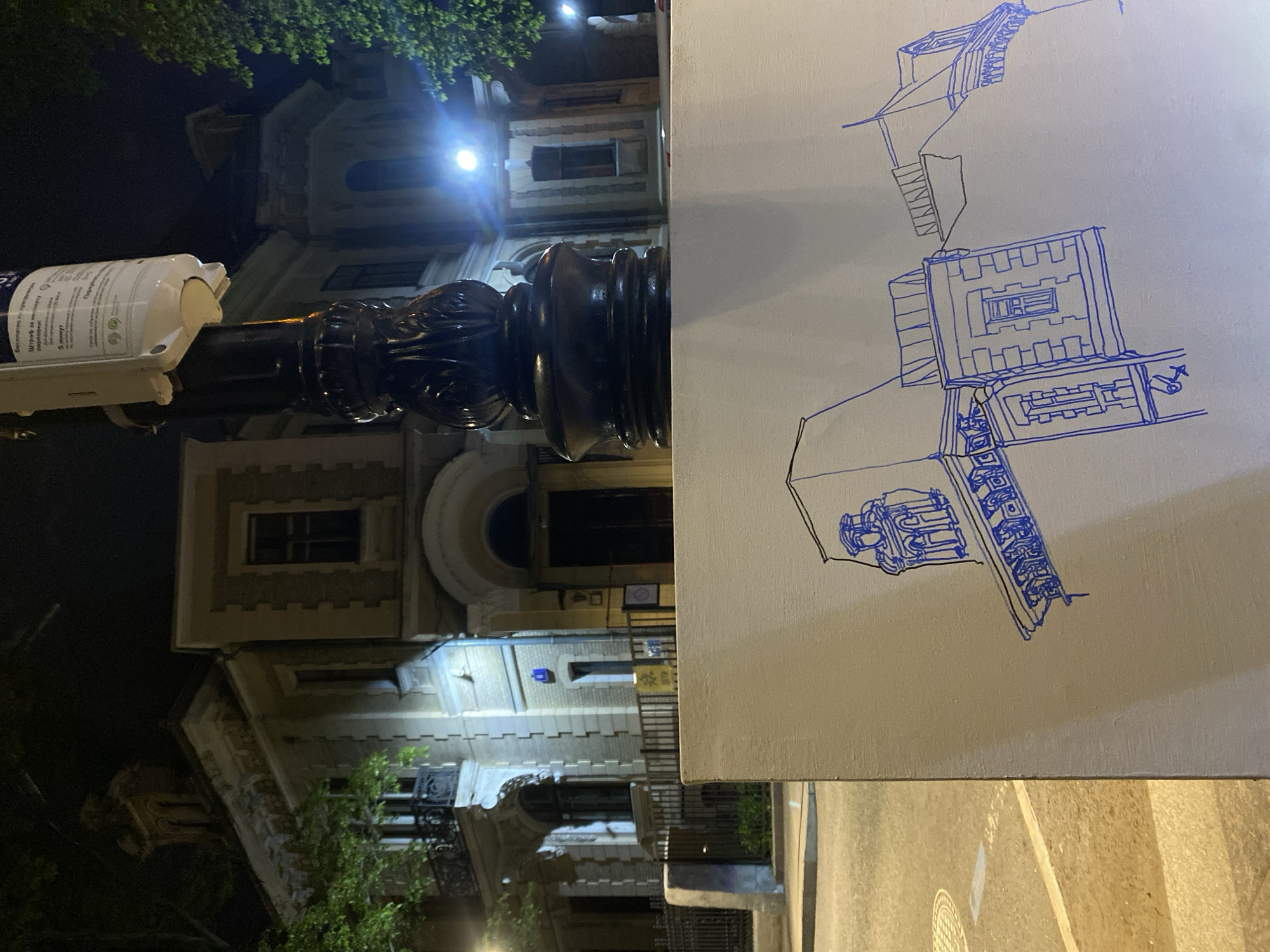 A street scene at night in Moscow, Russia, featuring a street lamp and a canvas leaning against it. The street lamp has a decorative black base and a white cylindrical attachment at the top. The canvas has a blue-ink drawing of an architectural structure. The background displays a building with lit windows and intricate architectural details, suggesting a historic or ornate design. The foreground shows a section of the street with asphalt and markings.