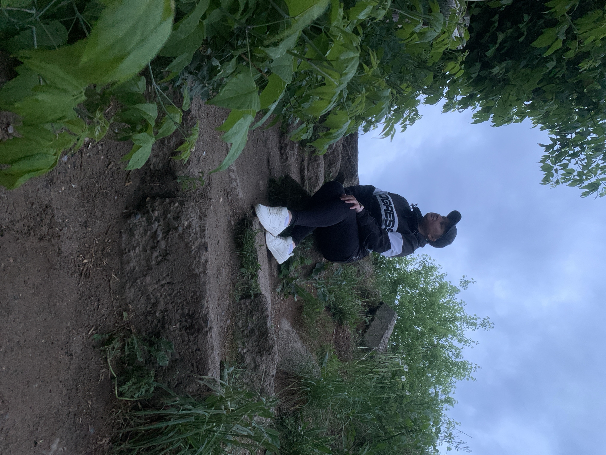 A person is sitting on a tiered, earthen structure covered in foliage, possibly a hillside or terraced landscape. The person is wearing a black jacket with white stripes on the sleeves, black pants, white sneakers, and a black cap. The person is facing towards the right side of the image, slightly angled upwards. The sky is overcast, and greenery including trees and plants frame the upper edges of the composition. The location of Chaikovsky, Russia, is unknown from the image content alone.