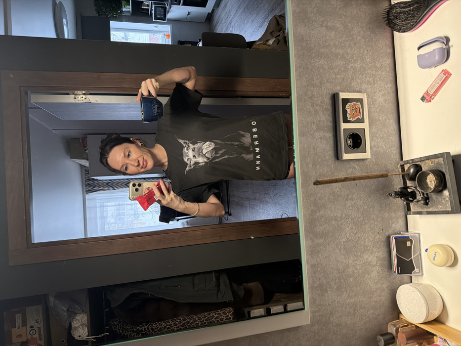 This image captures a female subject taking a mirror selfie in an apartment interior located in Perm, Russia. The adult female, with dark hair styled up, wears a black t-shirt displaying a grayscale Doberman dog with its tongue partially extended, and the mirrored text "NNAMREBO" (originally "OBERMANN") below the dog graphic. She holds a light-colored smartphone in her right hand, positioned for the selfie, with a small red object visible behind the phone. Her left hand holds a dark blue, textured mug. A thin, light-colored bracelet is visible on her right wrist.
The foreground, below the mirror, features a white countertop. Resting on it are: a grey stone-like tray holding an incense burner with a lit stick, a small Buddha statue, and a bowl containing ash; a clear plastic holder with several credit cards; a small beige oval container; various small bottles on a wooden tray; a pink translucent lighter; a light purple earbud case; and a black hairbrush with pink bristles. An electrical outlet, featuring a European-style socket with a cartoon sticker of an orange character on one side, is mounted on the textured grey wall above the countertop.
The background reflected in the mirror reveals an interior apartment space. To the left, an open closet or storage area contains hanging clothes, including a leopard-print garment, and stacked boxes on a shelf. Through a wooden door frame in the center-right, another room is visible, appearing to be a kitchen or living area. This room features a window with white curtains, light-colored kitchen cabinetry, a microwave, an oven, and shelves adorned with plants or decorative items. A dark grey wall section and a ceiling light fixture are also present. The flooring visible in the reflected room is wood or laminate.