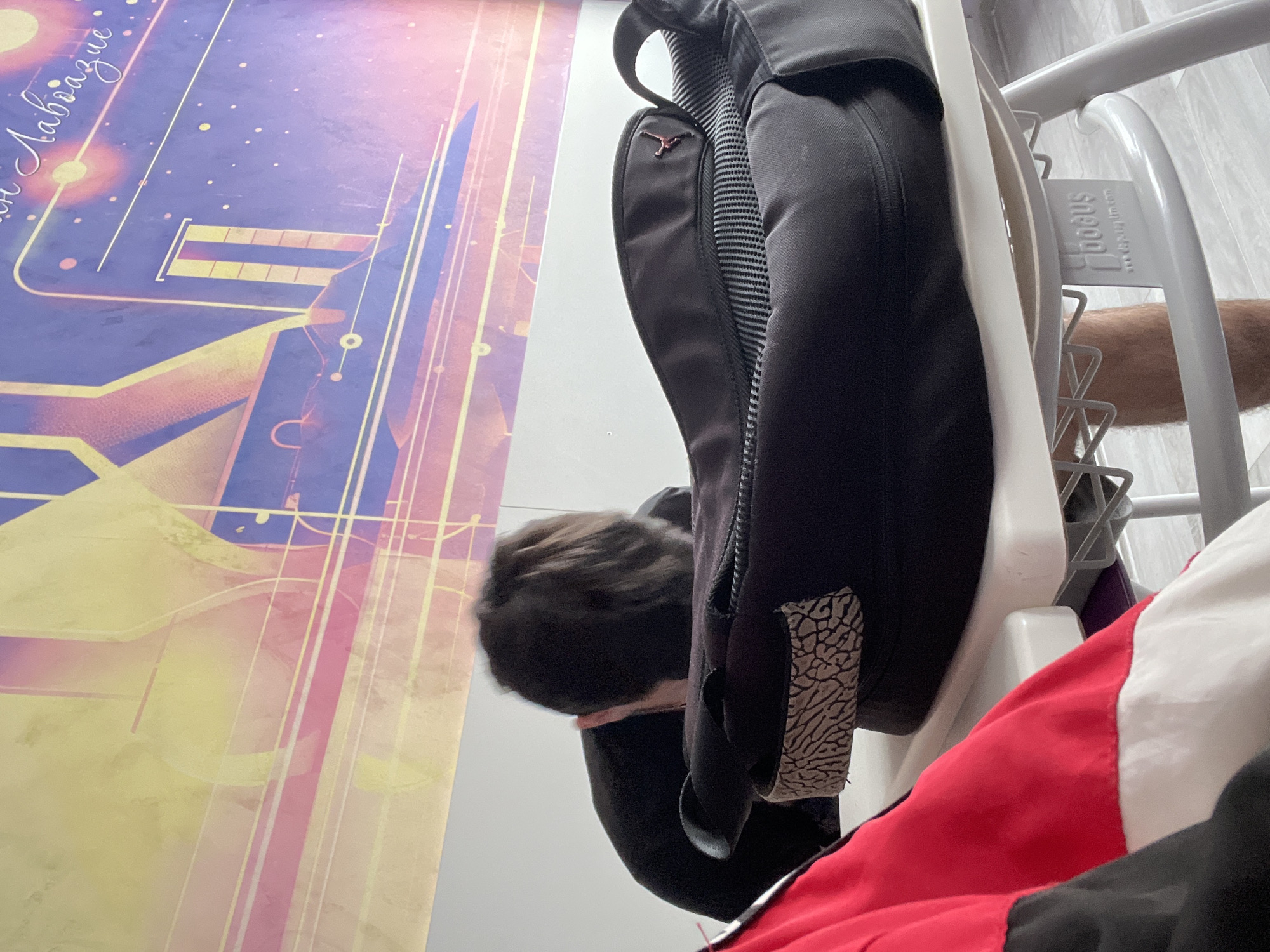 The image captures an indoor scene, likely a casual moment in a room. The primary subjects include a partially visible person, objects like a backpack and furniture, and a large poster on the wall.
To the left, a vibrant, abstract poster or piece of artwork adorns a wall, featuring a cosmic or futuristic design with lines, geometric shapes, and what appear to be stars or glowing celestial bodies. Predominant colors are shades of purple, yellow, and pink. Partially visible Cyrillic text, possibly "он Лавазище" (on Lavazishte), is inscribed on the poster.
In the foreground and center, a black backpack is prominently displayed, possibly hanging or resting on a white, tubular metal structure, which appears to be part of a chair, bed frame, or storage unit. A distinctive red "Jumpman" logo, associated with the Jordan brand, is visible on one of the backpack's straps. Another notable detail on the backpack is a grey elephant print pattern on one of its panels.
To the immediate right of the backpack, the back of a person's head with dark, short hair is visible, turned downwards. The person is wearing dark clothing. Further to the right, a multi-colored garment, primarily red and white with some black, is visible. Below this, a portion of a human arm is seen.
The white metal structure has a white plastic component where the brand name "Hogus" and the country "Bulgaria" are embossed, providing a specific location cue consistent with Plovdiv, Bulgaria. The activity depicted is ambiguous but suggests a moment of rest or casual interaction within an indoor setting. The lighting appears to be artificial indoor illumination.