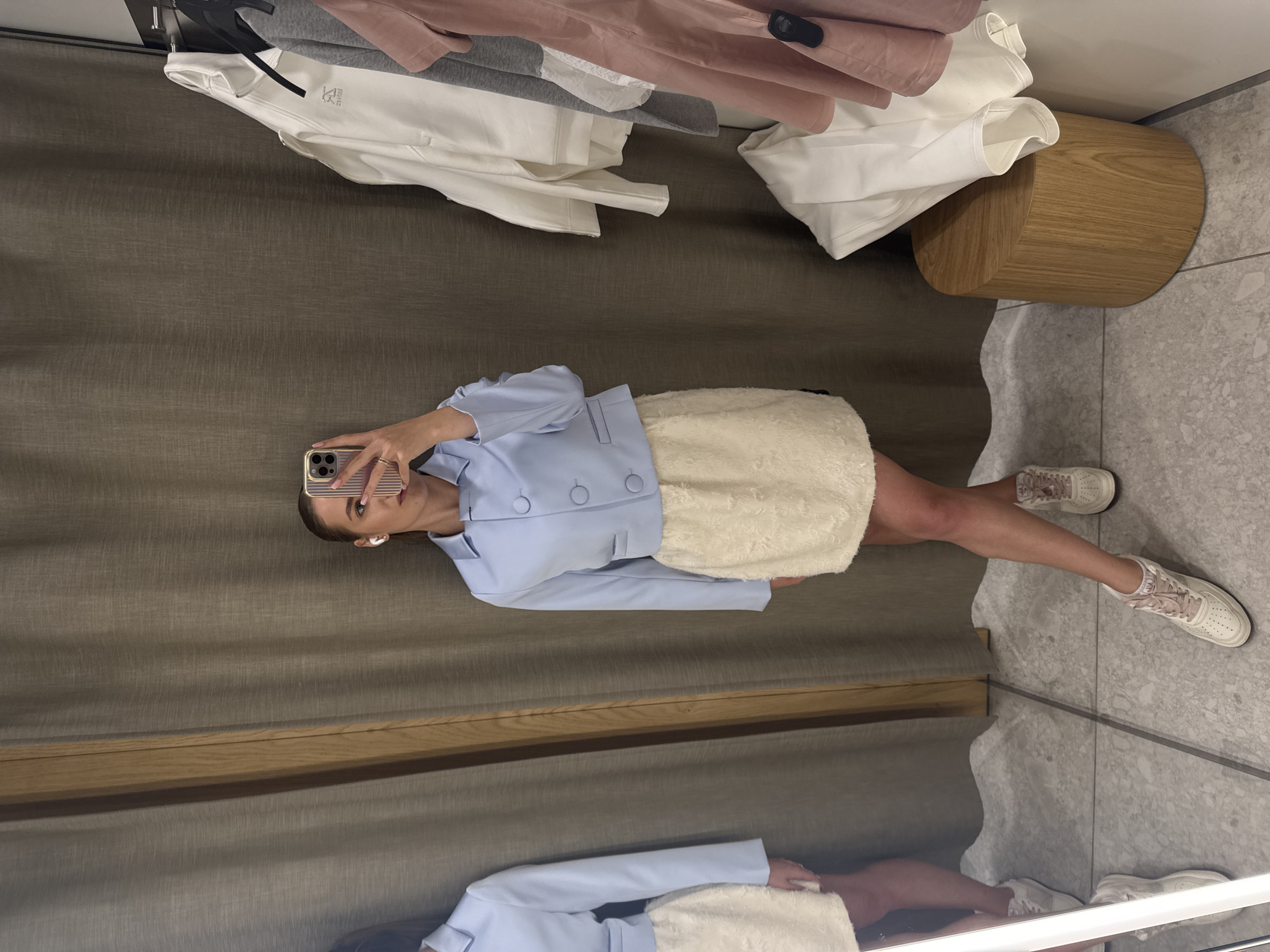 A woman stands in a fitting room, taking a mirror selfie with her smartphone. She is wearing a light blue cropped blazer over a cream-colored textured skirt and white sneakers with pink laces. She has her hair pulled back and is wearing white wireless earbuds. To her right, several garments, including a white zip-up jacket, a gray t-shirt, and a pink t-shirt, hang on a rack. A cylindrical wooden stool sits on the tiled floor to the right of the frame. The fitting room has grey curtains and a wooden framed mirror.