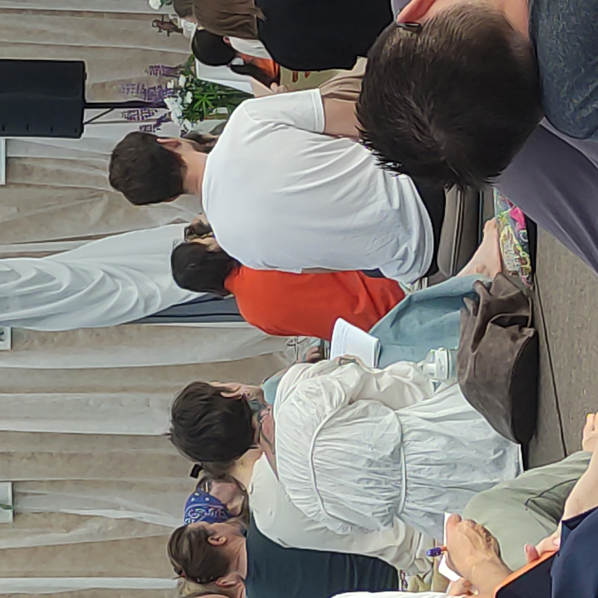 The image shows a group of people sitting on the floor, facing away from the camera. The setting appears to be an indoor event, possibly a workshop or a lecture, with draped fabric in the background and a speaker system visible. The people are dressed casually, and some are holding papers or writing. One person in the foreground is wearing a white top, and another is in an orange top. The lighting is soft and diffused. The image is a photograph taken from a slightly elevated angle, looking down at the seated individuals. There is no visible indication of the location being Zhirnosovo, Russia; this information is not supported by the visual evidence in the photograph.