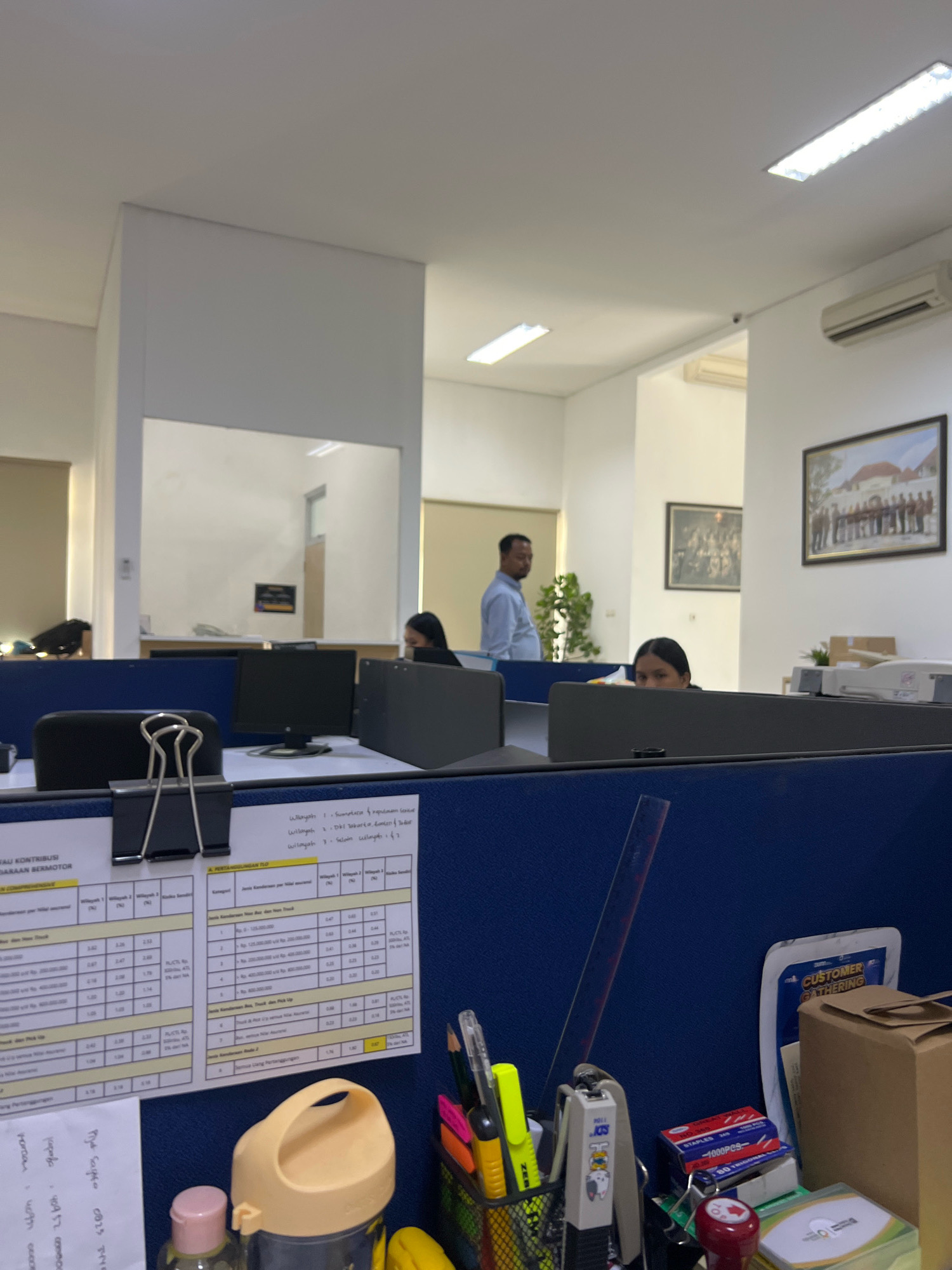 The image depicts an office environment, likely a cubicle farm, with several individuals present. In the foreground, a blue fabric partition separates cubicles. A large black binder clip is attached to a document on this partition. A black computer monitor and keyboard are visible on a desk. To the right, another blue partition is adorned with various documents, including charts and tables with Indonesian text and numbers. A ruler leans against this partition. On the desk area to the right, a stapler, pens in a black mesh holder, a yellow water bottle, and a box of staples are arranged. Further back, a woman with dark hair is seated at a desk, looking towards the camera. Behind her, another partition offers a glimpse into another office area, where a man in a light blue collared shirt is standing and looking away from the camera. The background shows white walls, fluorescent lighting, and framed pictures on the wall, including one depicting a group of people in formal attire and another with buildings. An air conditioning unit is mounted high on the wall. The overall scene suggests a typical office setting in Yogyakarta, Indonesia, likely a workspace for administrative or business operations.