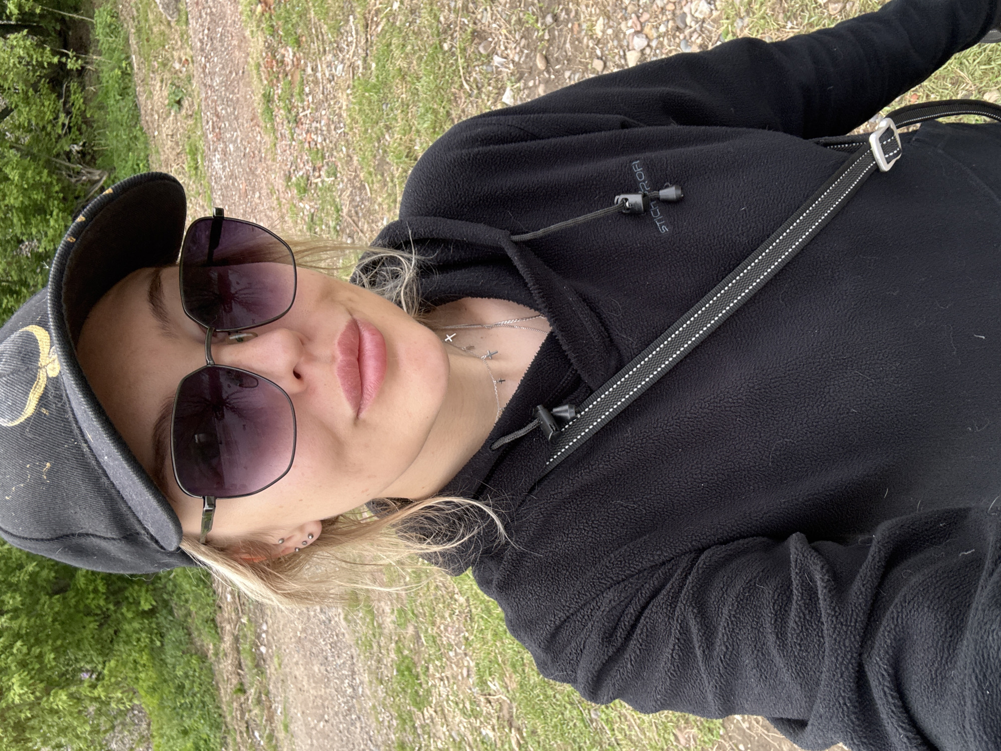 A woman is shown in a medium close-up shot, wearing a black baseball cap with an indistinct gold design, sunglasses with dark lenses, and a black fleece hoodie. A black strap with white detailing runs across her chest. She has blonde hair and multiple piercings in her left ear. A delicate silver necklace with cross pendants is visible. The background is a blurred mix of green grass, trees, and a dirt path.
The location is ambiguous and I cannot confidently state that this image was taken in the city of Krasnoyarsk, Russia.