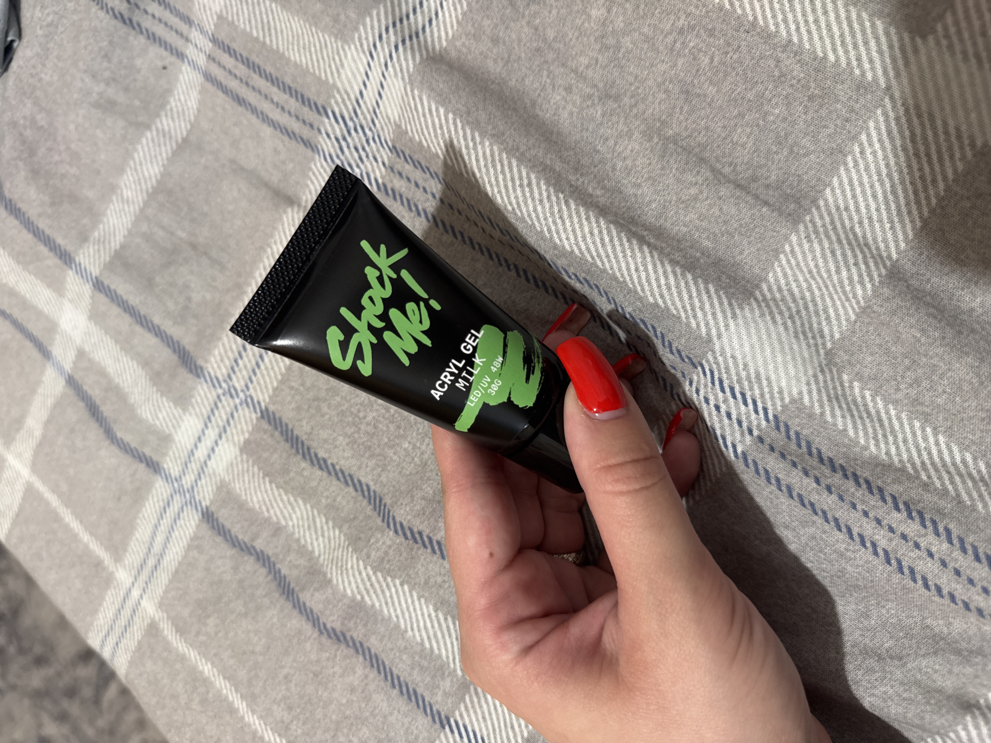 A hand with bright red nail polish holds a black tube of "Shock Me!" acrylic gel. The tube has green lettering and a black screw-top cap. Text on the tube reads "ACRYL GEL MILK" and "LED/UV 48W 30G." The hand and tube are positioned on a grey plaid blanket. The image is a close-up shot, focusing on the product and the hand holding it. The environment appears to be an indoor setting, with soft lighting. There is no indication of the city of Яромінскі сельскі Савет, Belarus.