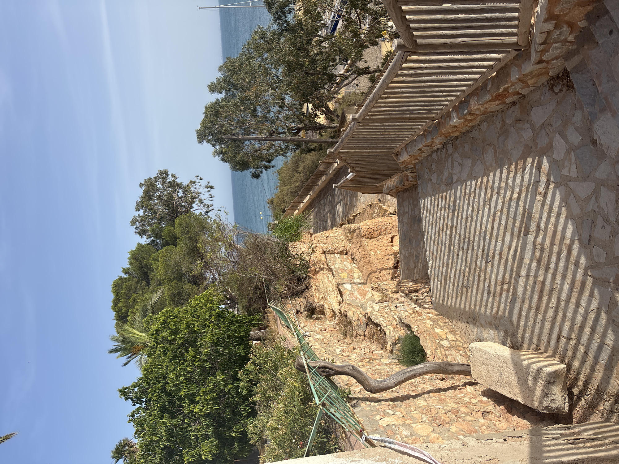 This is a photograph taken in Urbanización Cabo Roig, Spain, showing a stone pathway descending towards the sea. The pathway is bordered by a rustic wooden fence on the right and a rough-hewn stone wall on the left, which supports a sloped garden area with various trees and bushes. The stone steps are irregular in shape and size, creating a natural and textured surface. Sunlight casts strong, linear shadows across the steps, indicating a bright, sunny day. In the background, the turquoise sea is visible, with a clear blue sky above. A few boats, including a sailboat mast, can be seen docked in a harbor to the right, and a distant shoreline is visible on the horizon. There are no people visible in the image. The overall scene depicts a scenic coastal walkway, likely a path leading to a beach or marina, in a Mediterranean setting.