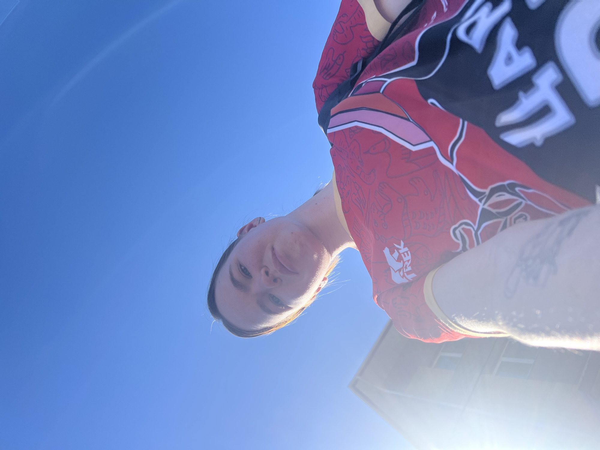 The image shows a person, likely female, from a low angle looking up at them. The background is a clear blue sky, suggesting daytime. Part of a building is visible in the lower right corner, partially obscured by glare from the sun. The person is wearing a colorful jersey-like garment with writing. The writing appears to be "TREK" and other text in a foreign script, possibly Cyrillic. The location is unknown, and I do not have enough information to determine if it is Силикатный, Russia.