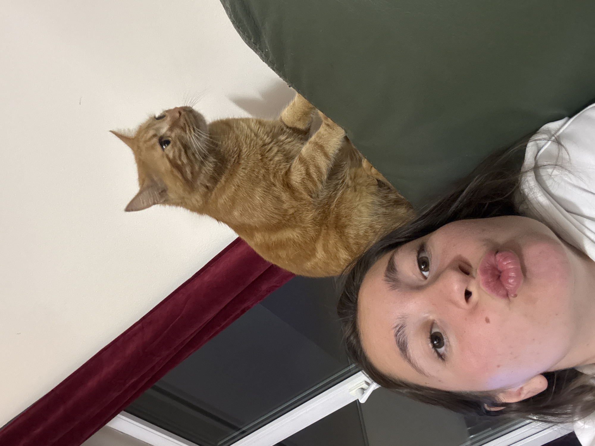 An orange tabby cat sits perched on a dark green cushioned surface, looking to the right of the frame. In the foreground, a young woman with dark hair and brown eyes makes a kissing gesture with her lips, her face angled towards the camera. She wears a white hooded garment. Behind the cat, a dark red curtain is visible, draped over a window with white frames. The background wall is off-white. The scene appears to be indoors, likely in a residential setting, possibly in Sofia, Bulgaria.