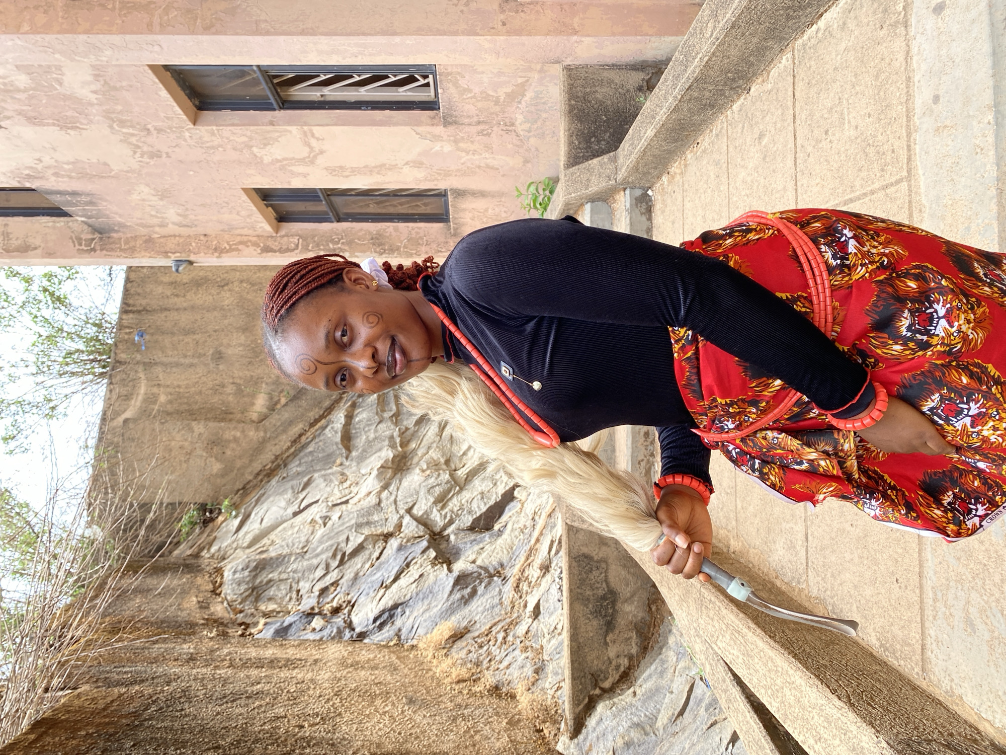 A woman stands outside a building in Gwong, Nigeria. The woman has brown braids, dark skin, and black face paint. She wears a black long-sleeved shirt and a red skirt with a lion pattern. She holds a ceremonial whisk. The building behind her has multiple windows, and the background is a large rock formation.
