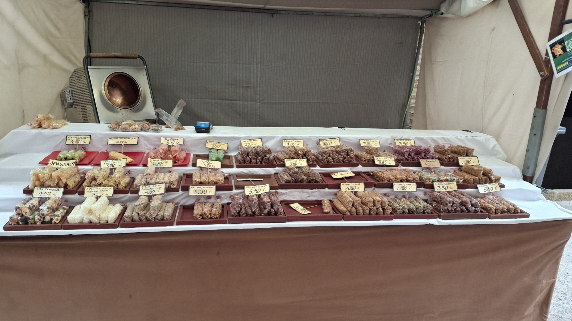This is a medium shot, eye-level, outdoor photograph of a food stall in La Fresneda / la Freixneda, Spain. The stall is covered by a white canvas awning. In the foreground, a brown tablecloth covers a table laden with various packaged food items. These items are displayed in small brown trays and consist primarily of nuts and dried fruits, with some jelly-like candies. Each item has a label indicating its name and price in Euros.

The display is organized in rows on tiered levels. On the top tier, there are several packages of what appear to be candied citrus peel or fruit slices, priced at 4.00 €. Next to these are bags of nuts, some with prices like 4.00 €, 4.99 €, and 3.50 €.

The middle tier features a variety of products. There are small, clear plastic bags containing what look like dehydrated fruits or vegetables, such as green slices labeled "JENGIBRE 3 €", and golden, irregularly shaped pieces. Other items include small, cylindrical rolls of nuts and dried fruit, some with labels like "COCO REHIDRATADO DESFIBRADO 3.50 €", "STICK DE JULI / GRANO 3.50 €", and "AVELLANA / HAZELNUT 4.00 €".

The bottom tier has more rows of similar packaged food items, including nuts, candies, and possibly small fruit bars. Prices visible include 3.50 € and 4.00 €.

In the background, on the top surface of the stall, a large, metallic pot with a wooden handle, possibly a candy-making machine, is positioned to the left. Next to it are several small bags of nuts or seeds. A small, dark blue electronic device, possibly a cash register or scanner, sits near the center. The stall is set against a large, gray corrugated surface. The overall impression is a well-stocked market stall offering a variety of confectionery and dried fruit products.