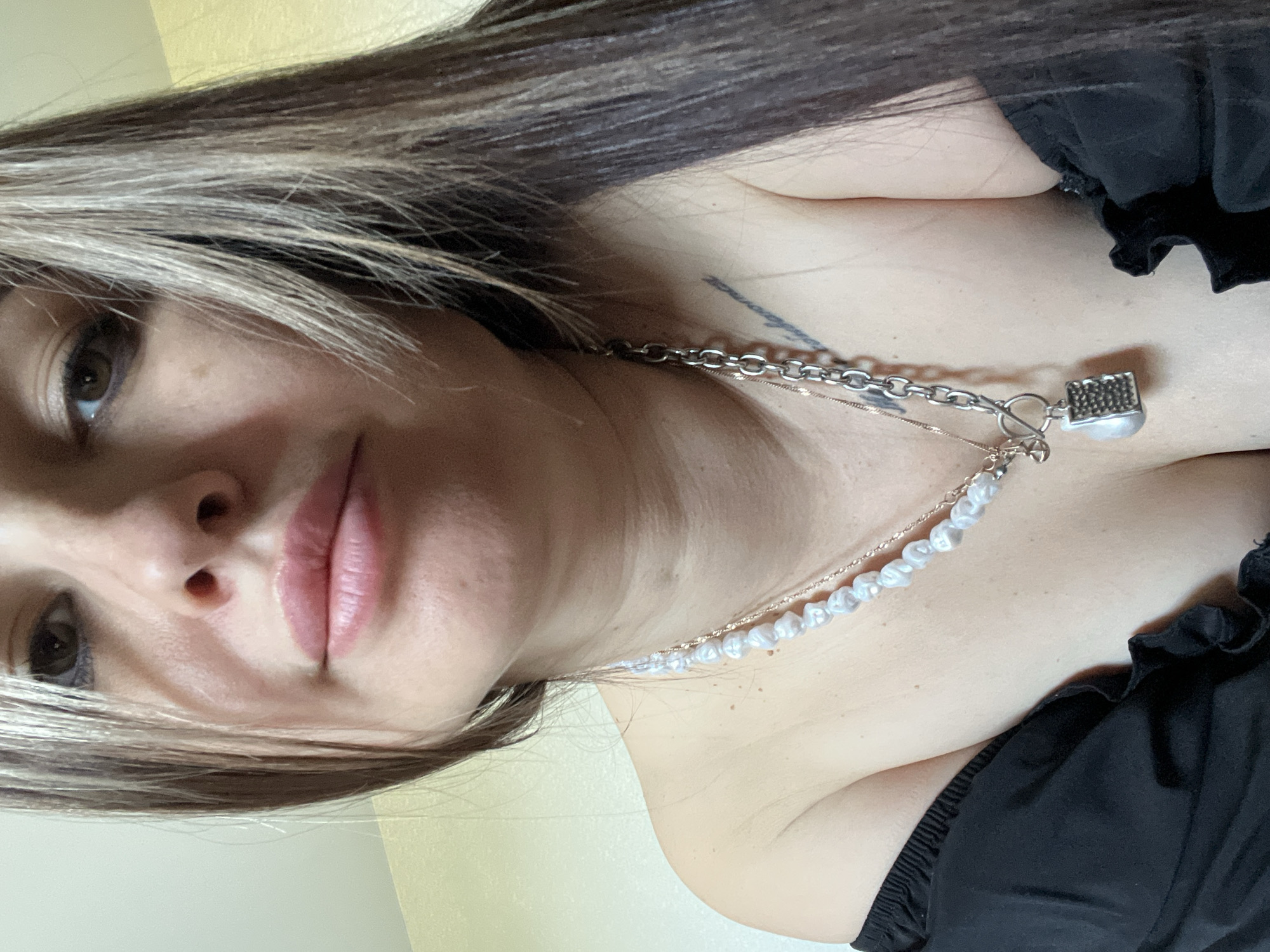 Here is a detailed description of the image:
A woman with long, dark hair and light brown highlights is the subject of this close-up portrait. Her face is turned slightly to the left, and her gaze is directed towards the viewer. She has light skin, defined eyebrows, and her lips are full and pink.
The woman is wearing a black, off-the-shoulder top. Around her neck, she is wearing a multi-layered necklace. The necklace consists of a silver chain, a strand of pearls, and a decorative pendant that appears to be a small padlock.
The background is a light yellow wall. The photograph is taken at an angle, giving a slightly tilted perspective.